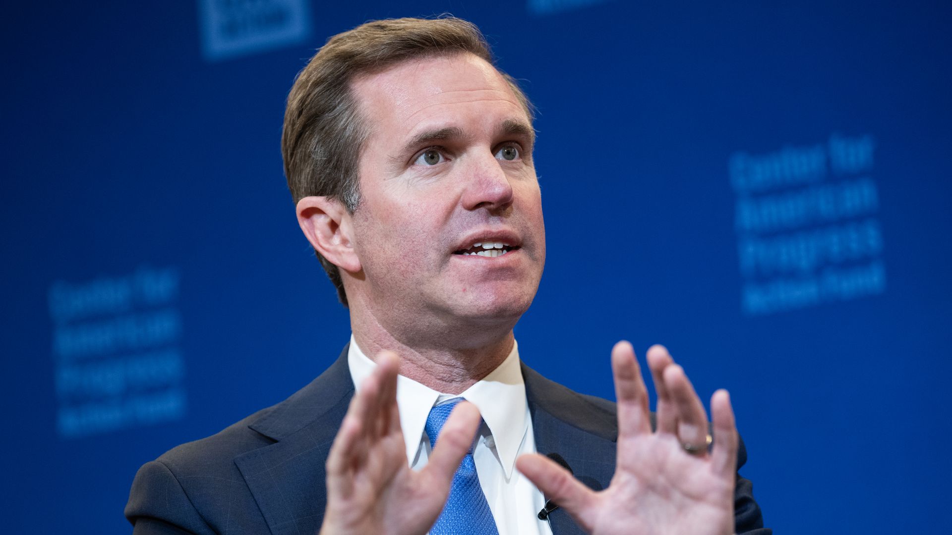 Kentucky Gov. Andy Beshear gestures with this hands, wearing a dark suit, white shirt and blue tie.