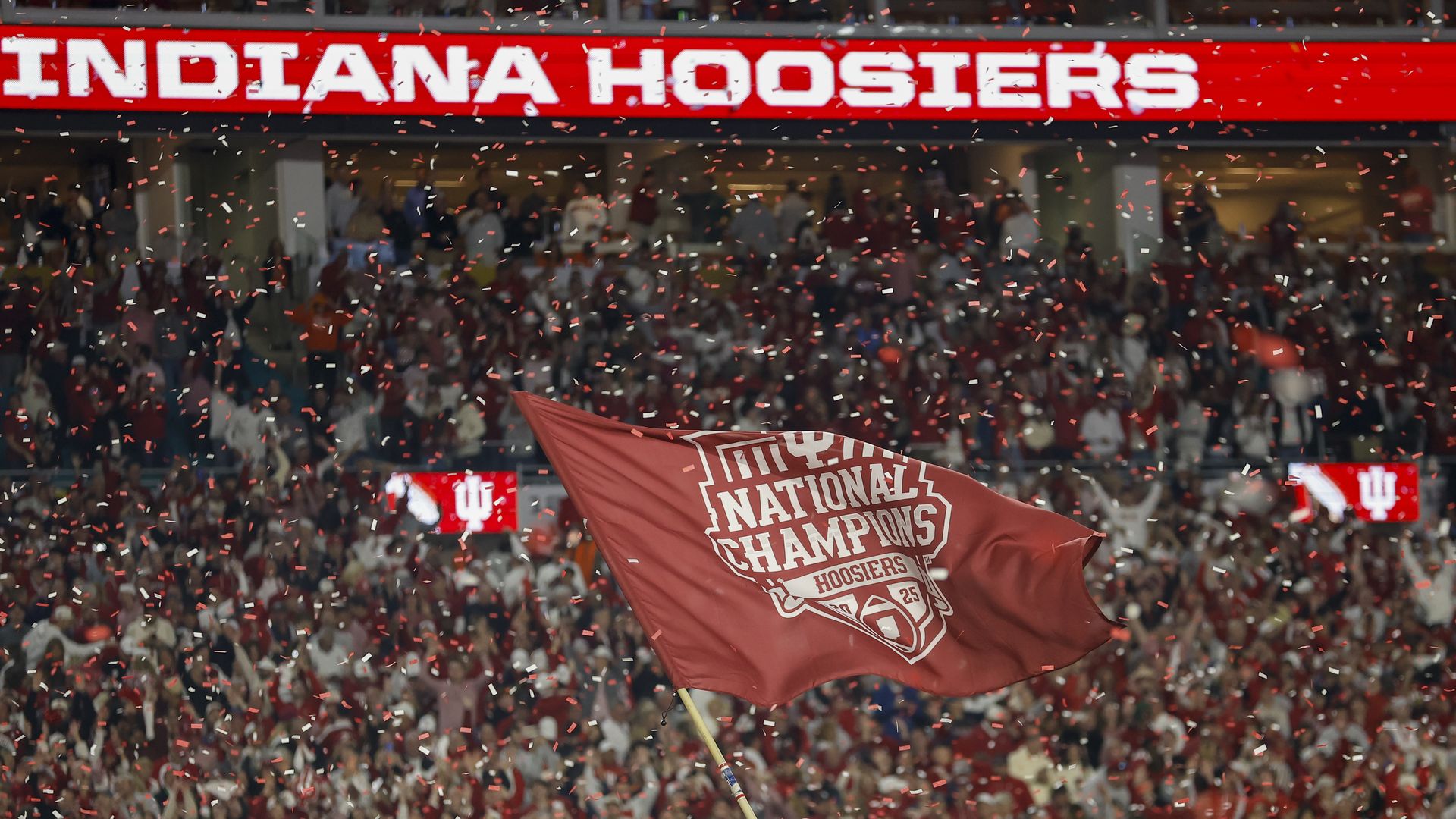 Red and white confetti fills the air over a crowd of fans in a stadium beneath a sign reading "INDIANA HOOSIERS" and a red flag waving that says "NATIONAL CHAMPIONS HOOSIERS".
