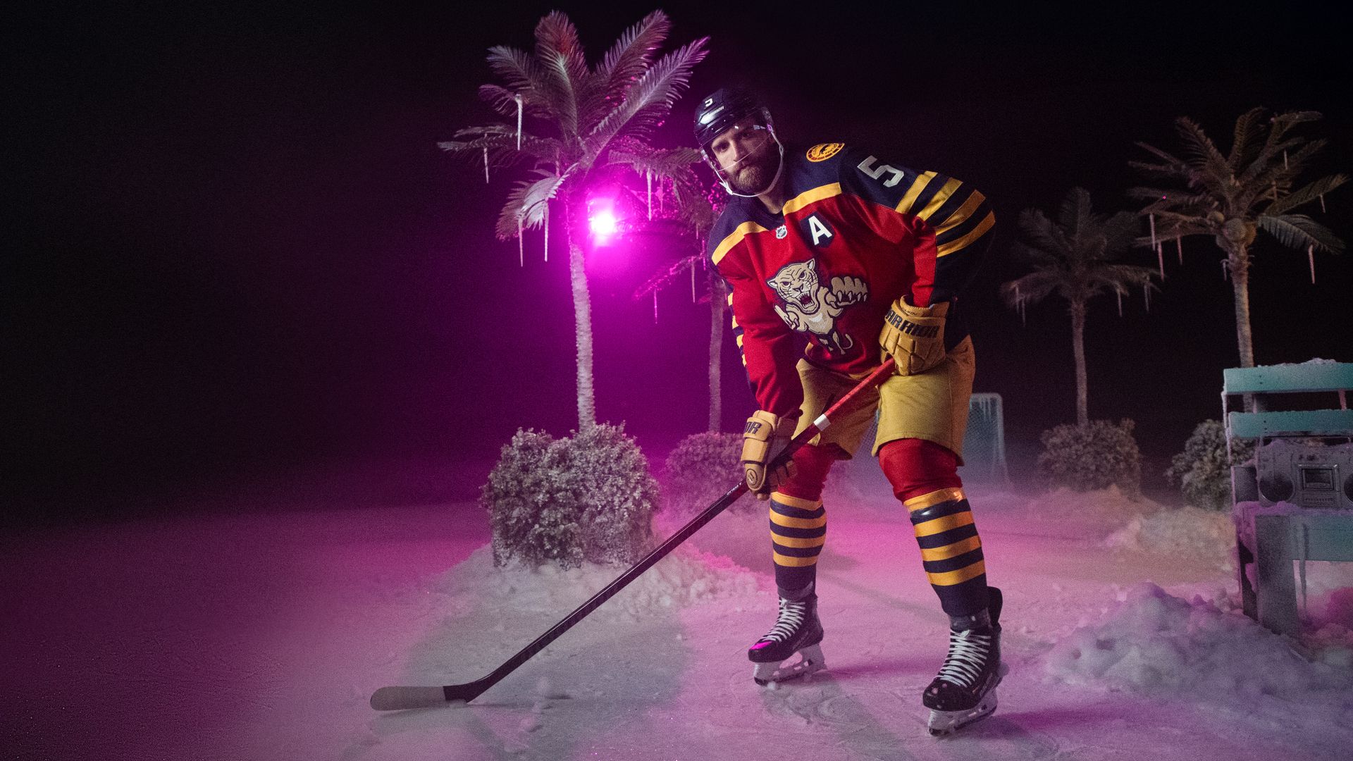 Ice hockey player in red, yellow, and black uniform posing with stick on ice rink at night, with palm trees and purple lighting in the background.