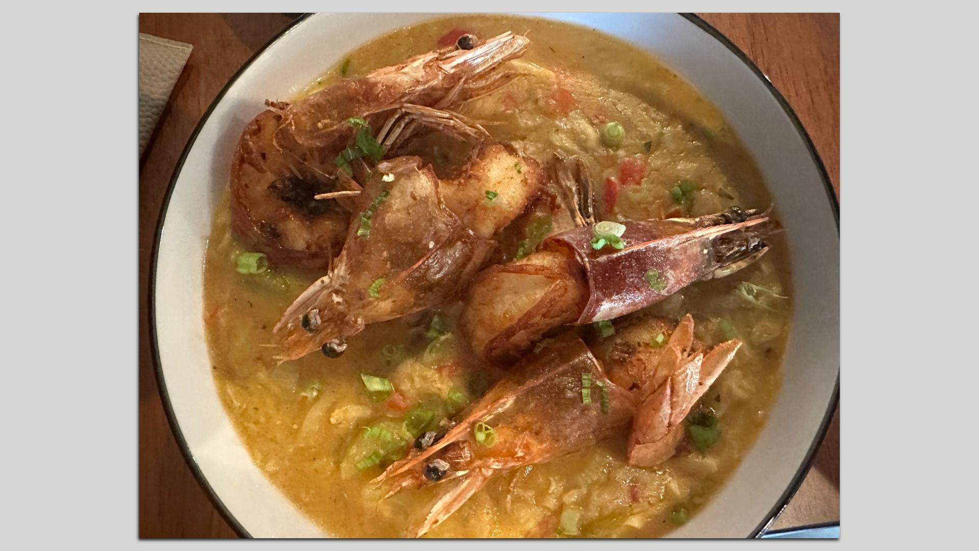 Creamy yellow shrimp soup in a white bowl with a dark rim, topped with large shell-on shrimp and sprinkled chopped green onions.