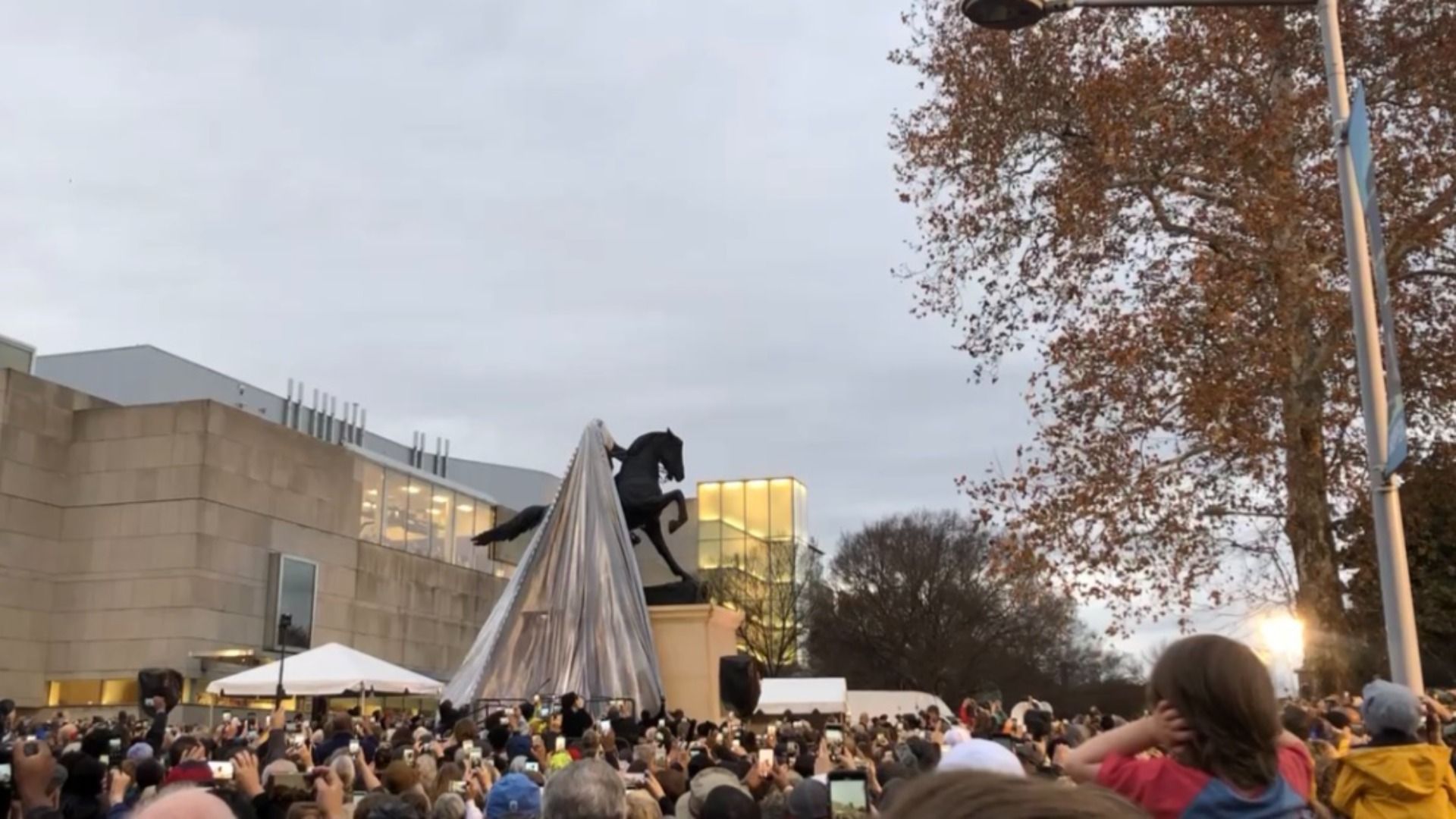 Crowd gathering at an outdoor plaza around a tall, triangular metallic sculpture with a black rearing horse on top; beige buildings, bare trees, and a cloudy sky.