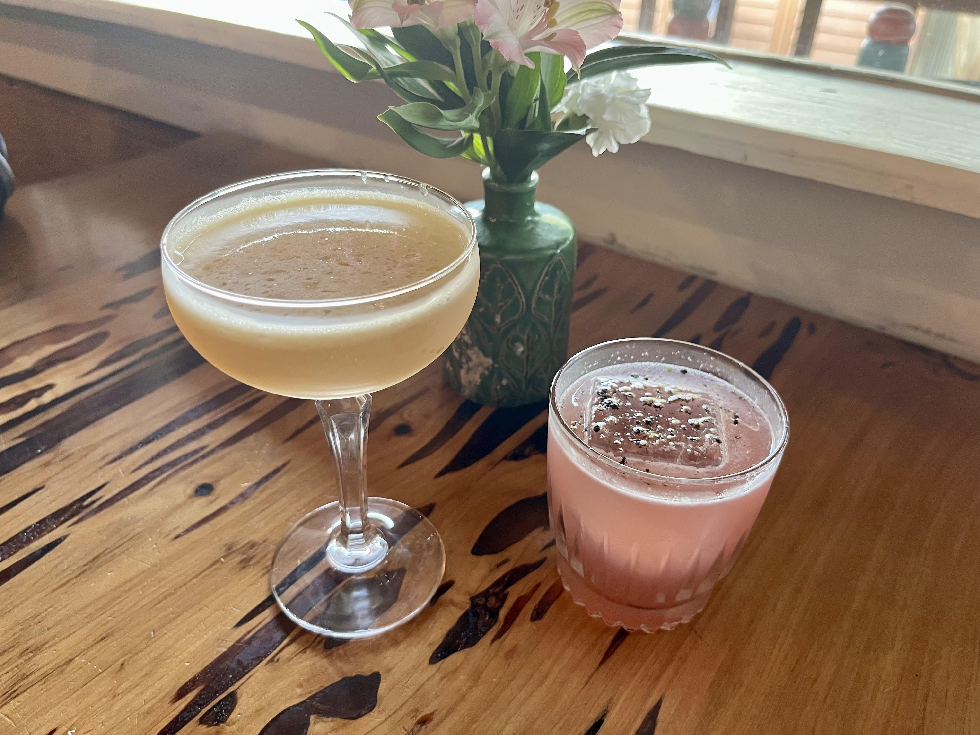 Two cocktails on a natural wood table with a flower vase and sunlight streaming in.