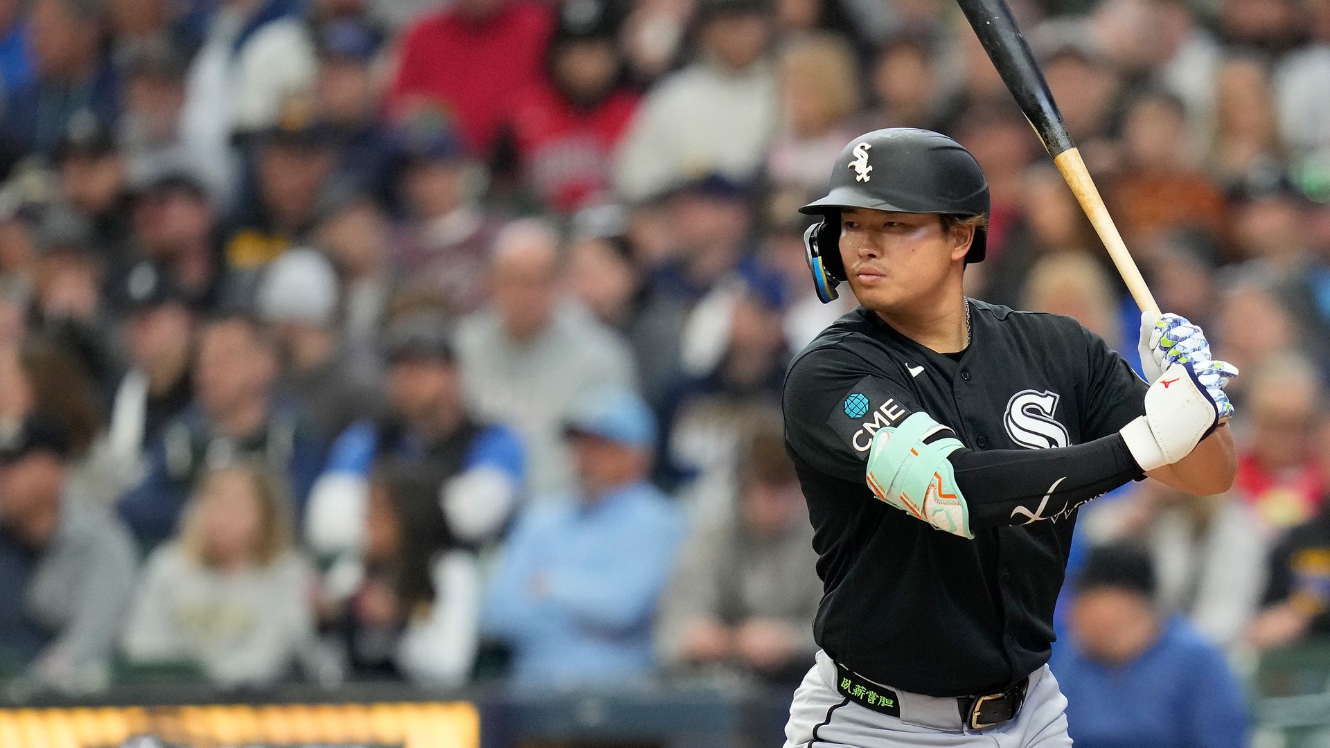 Baseball batter in a black Chicago White Sox uniform and helmet swings at the plate; turquoise forearm guard and white glove, with a blurred crowd in the stands behind.