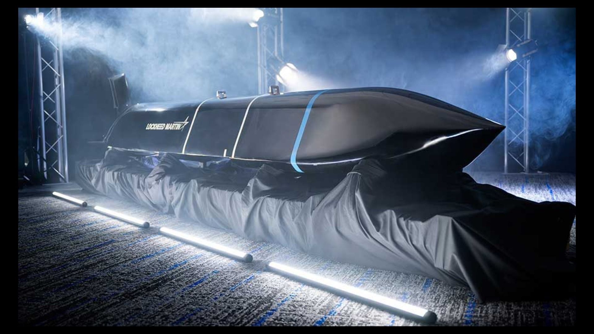 Sleek black missile with "Lockheed Martin" logo, covered partially by black fabric, displayed under bright white lights and surrounded by mist in a dark room.