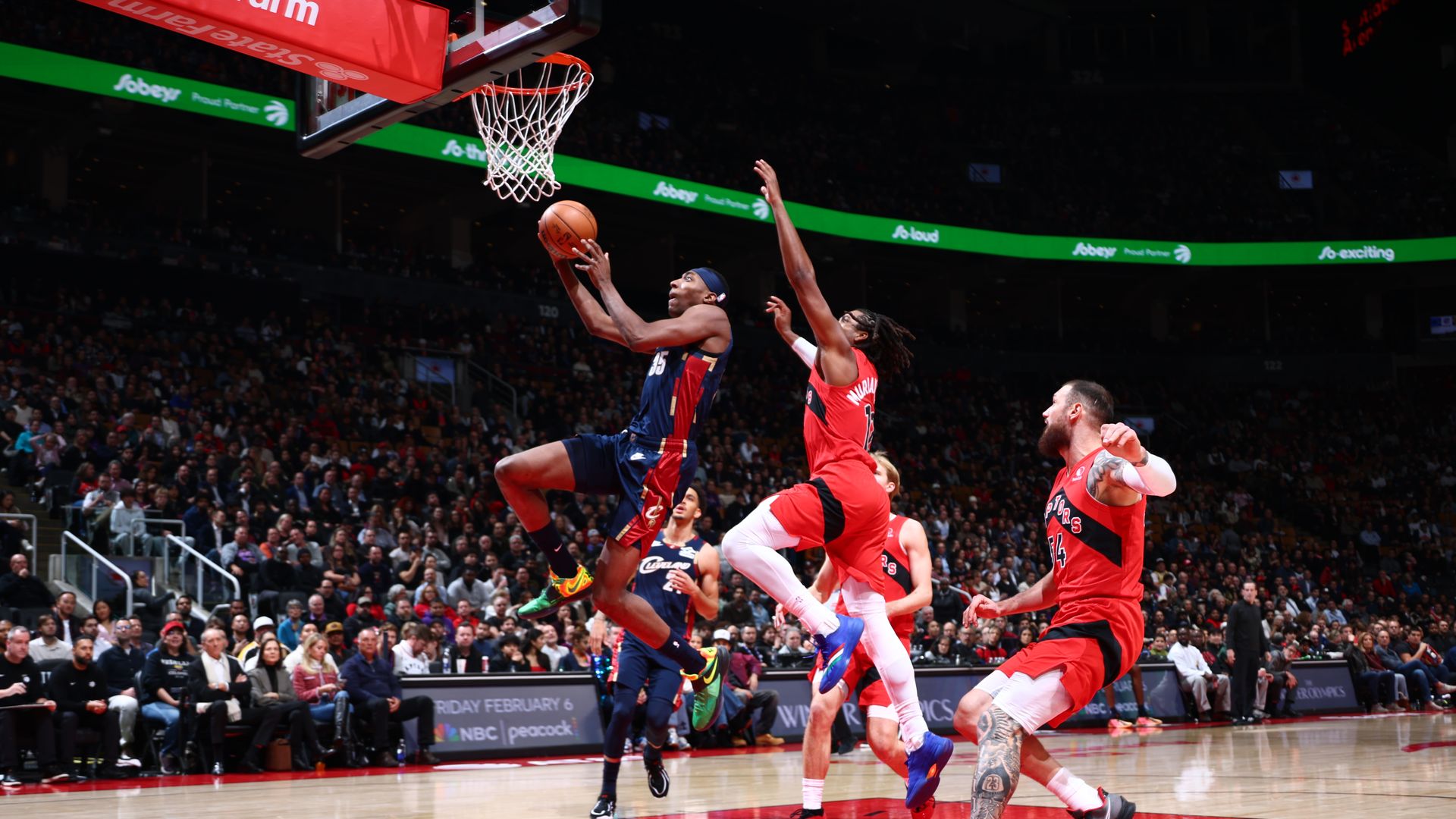 Basketball player in blue jersey drives to net, hounded by leaping player in red jersey. 