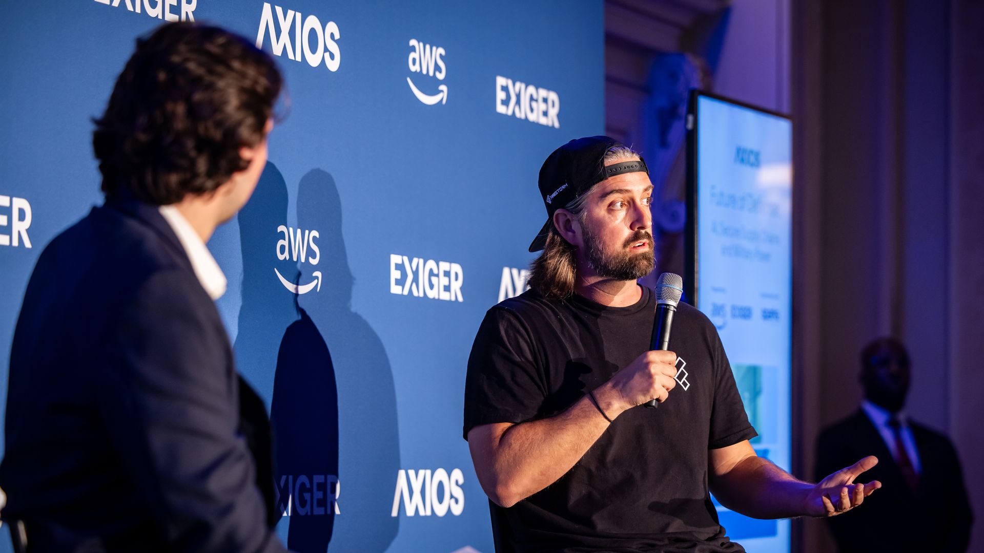 Two men sit on stage. The backdrop is blue with white lettering. A man stands offstage in a suit, to the right.