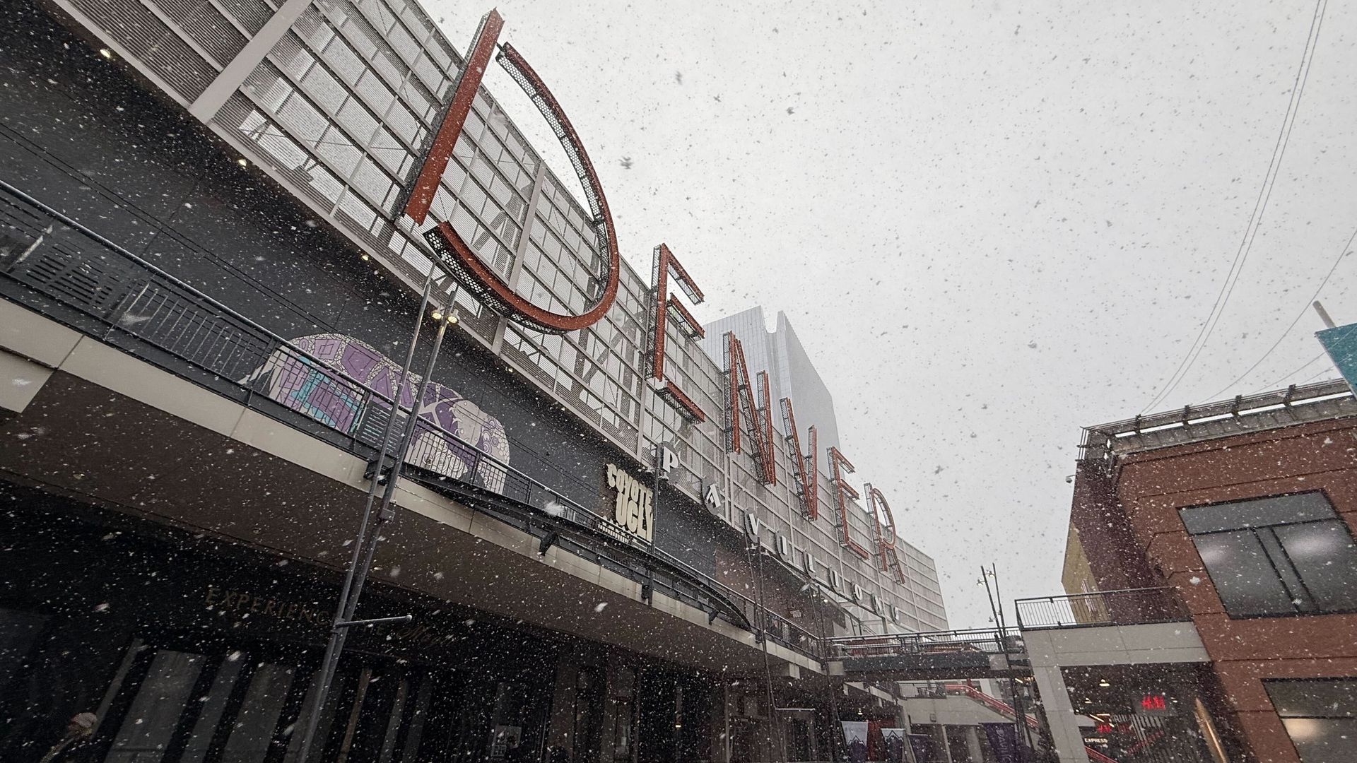 Snow falls over a modern, multi-level shopping center seen from below, with glass facades and large red-outlined letters; a brick building and a pedestrian bridge are visible.