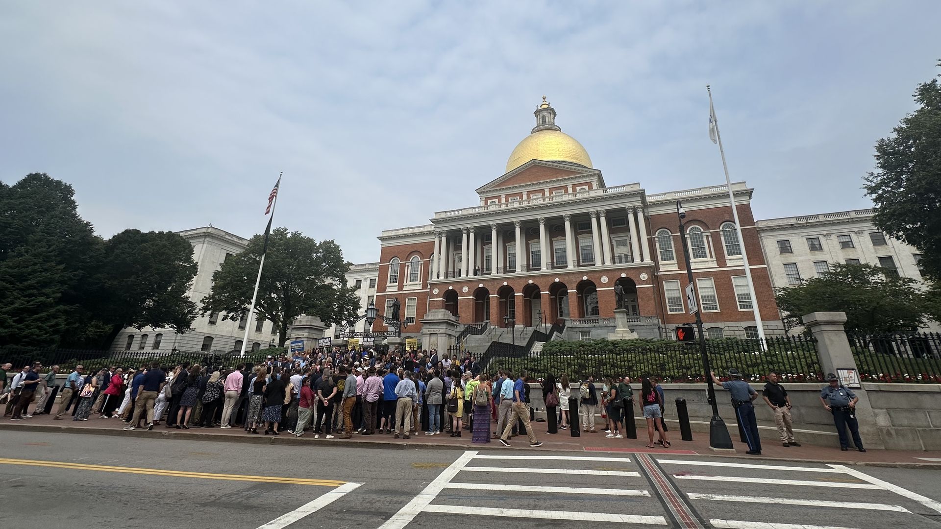 Crowd gathered outside large brick building with white columns and a gold dome under a cloudy sky, with American flag and crosswalk in foreground.