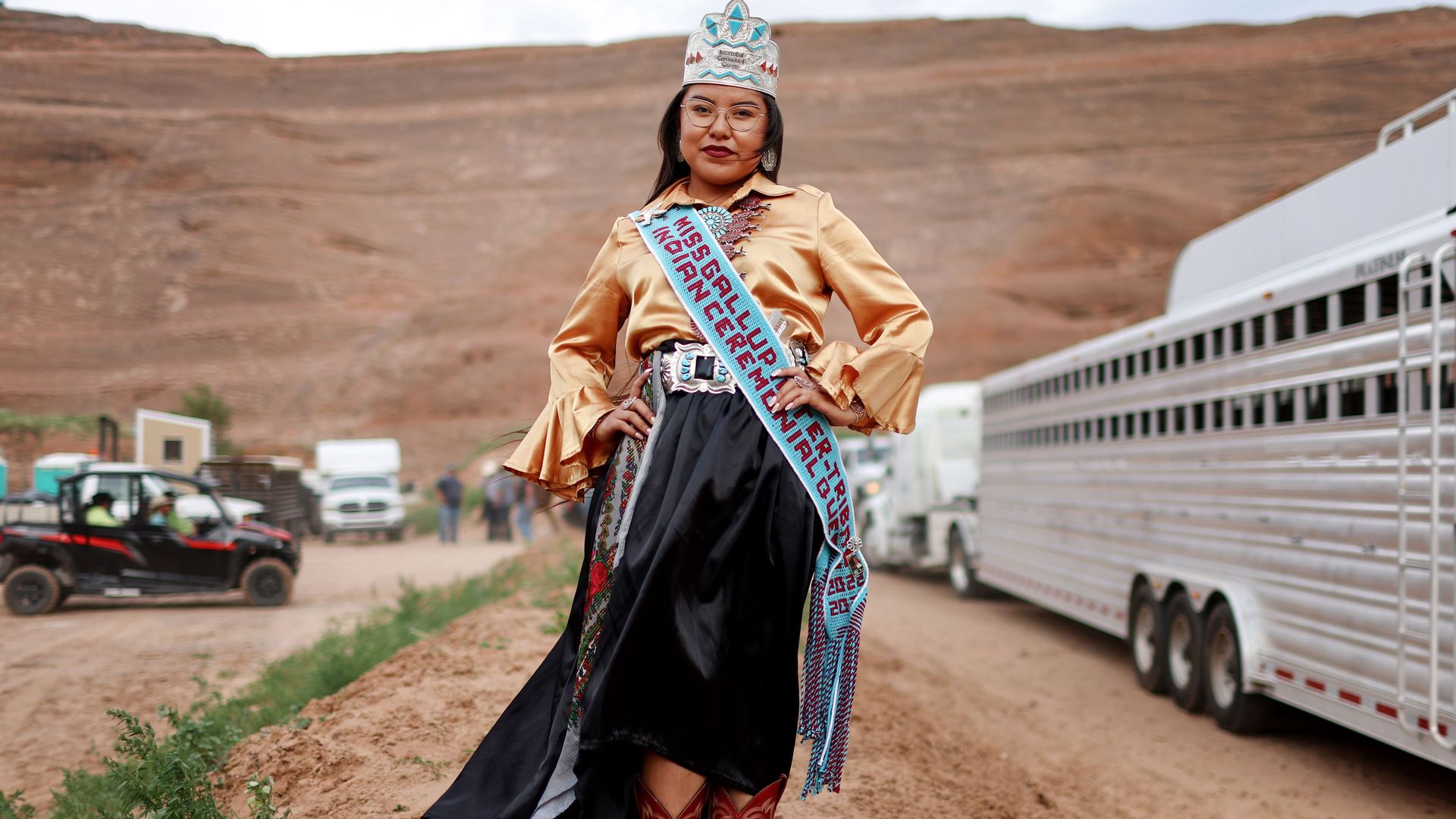 An indigenous woman from New Mexico stands with the mountains in the background. She is wearing a crown and sash
