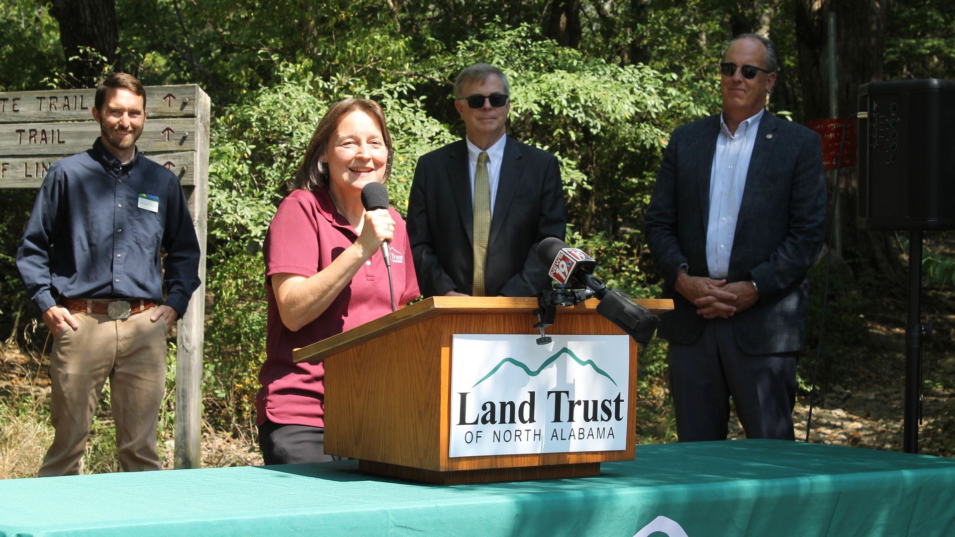 Woman in maroon shirt speaking at a Land Trust of North Alabama podium outdoors, with three men standing behind her near a forest trail sign.