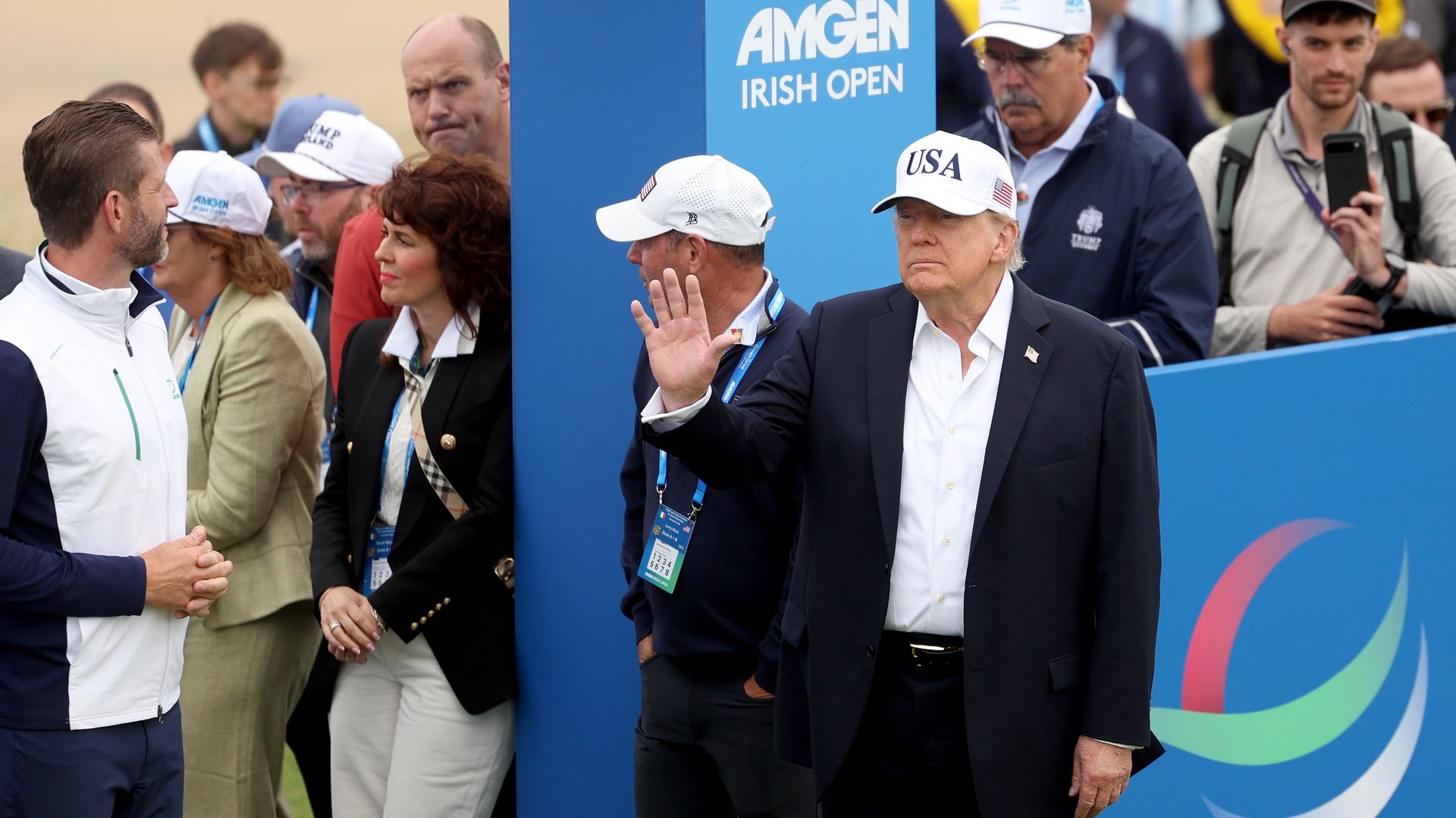 President Trump in a suit wearing a white USA cap waves to photographers at the Amgen Irish Open, beside a blue barrier, with officials and spectators in the background.