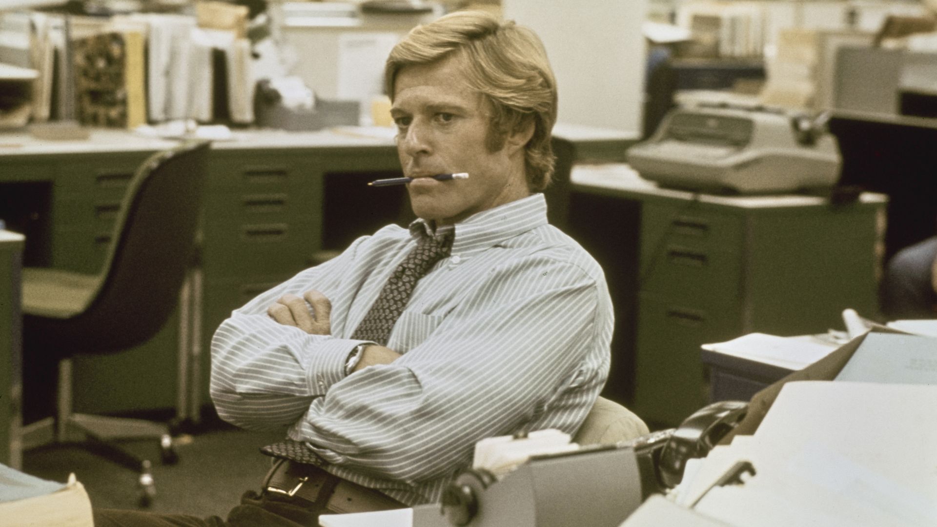 Blond man in striped shirt and tie sits in cluttered office with crossed arms and pencil in mouth, looking thoughtful amid desks, papers, and office equipment.