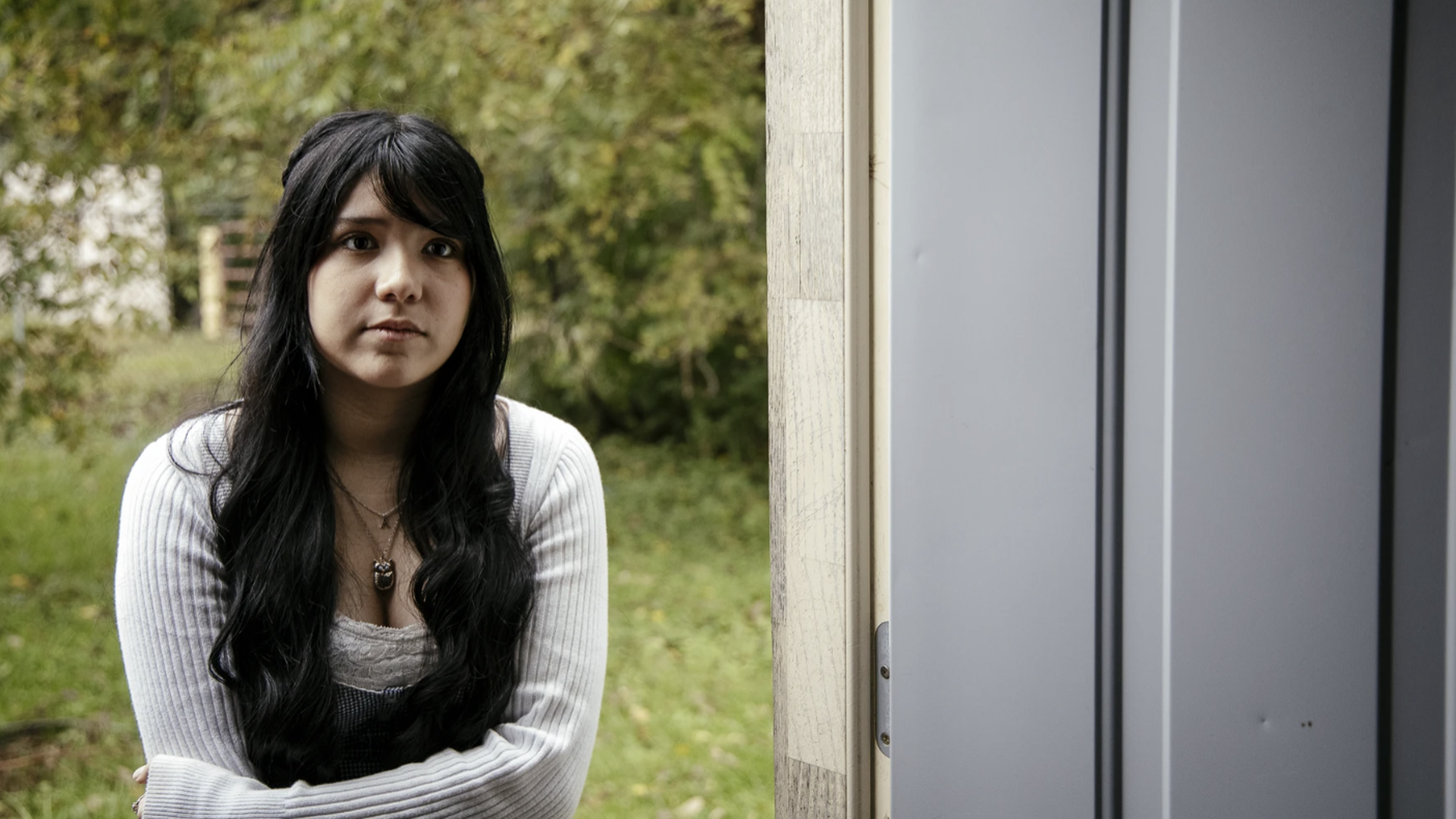 Portrait of a young woman with long black hair, wearing a white knit cardigan over a gray top and a pendant necklace, standing outdoors beside a light door frame with a green, leafy background.