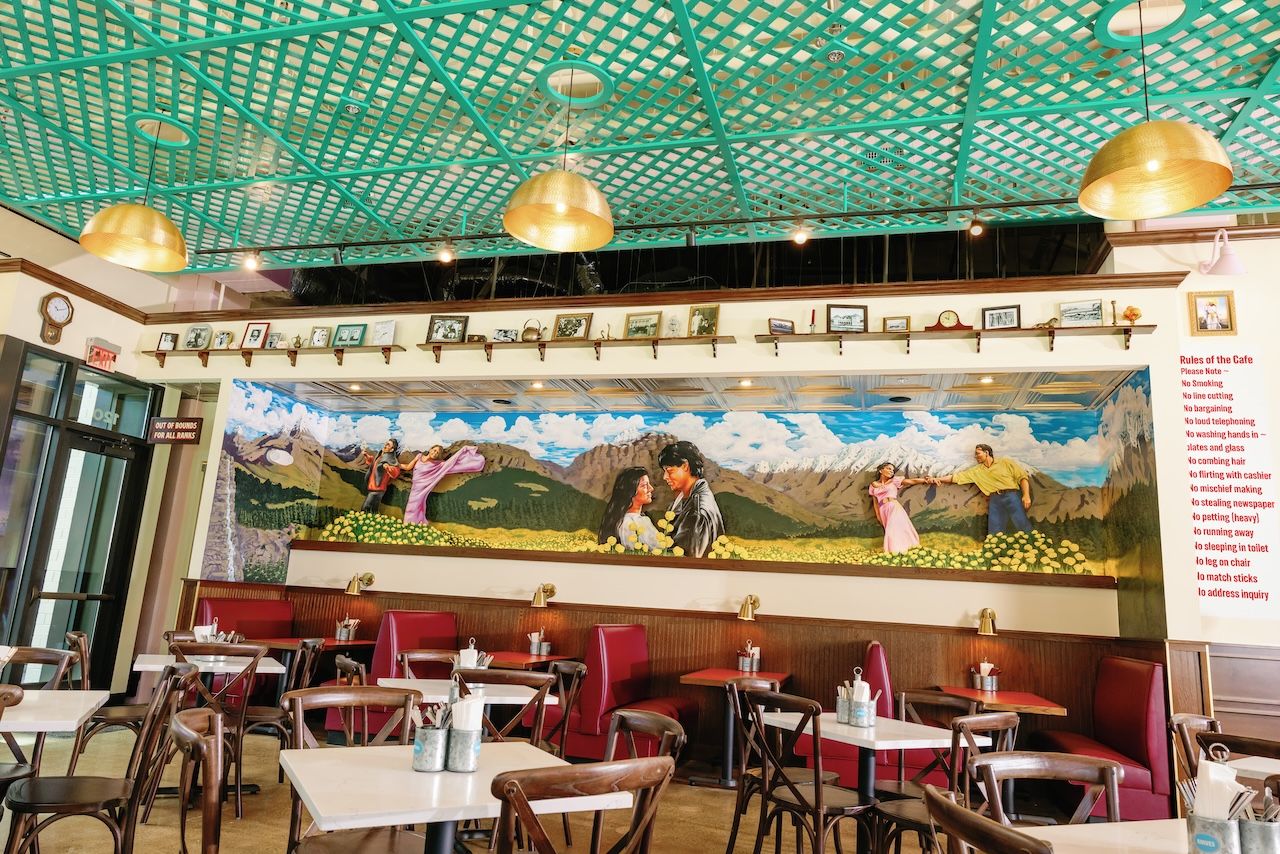Cozy cafe interior with a turquoise lattice ceiling and large gold pendant lights. A mural of mountains and people spans the wall, with red booths, white tables, and framed photos above.