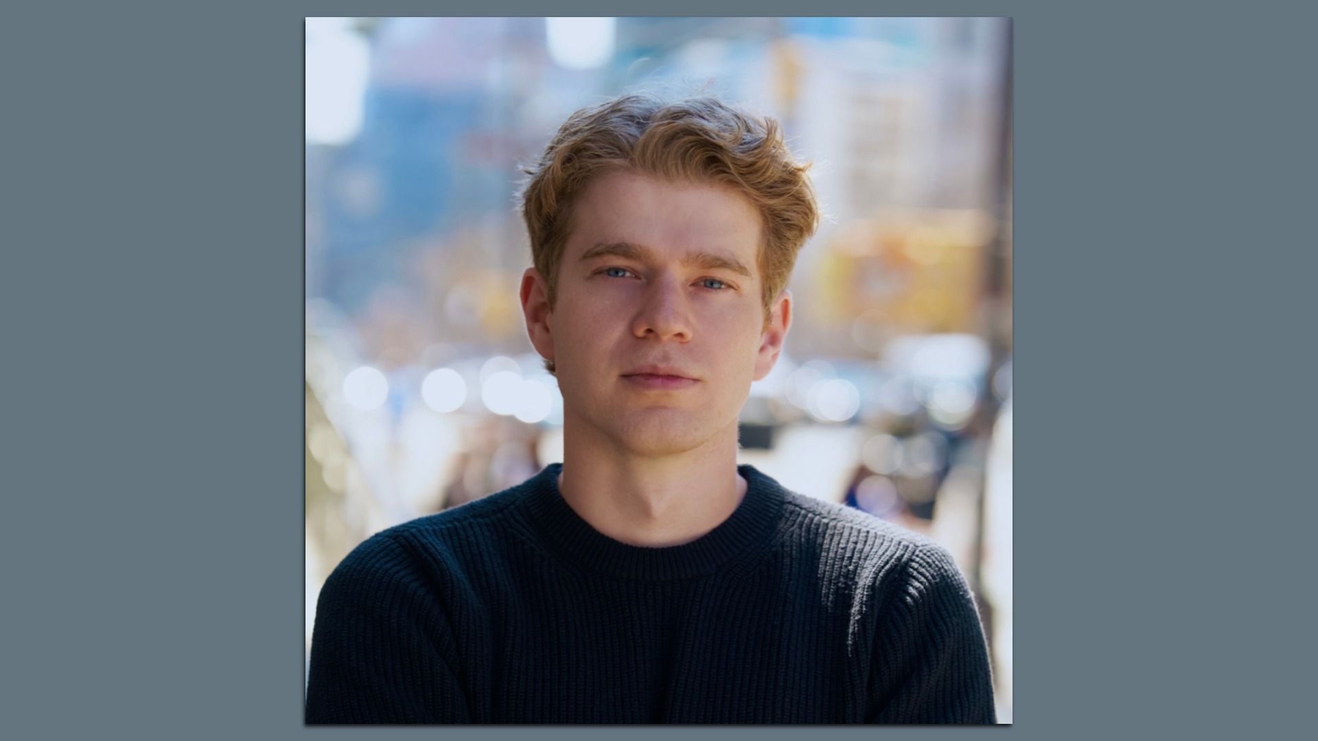 Portrait of a young man with blond hair and blue eyes, wearing a dark knit sweater, outdoors in a city with blurred lights in the background.