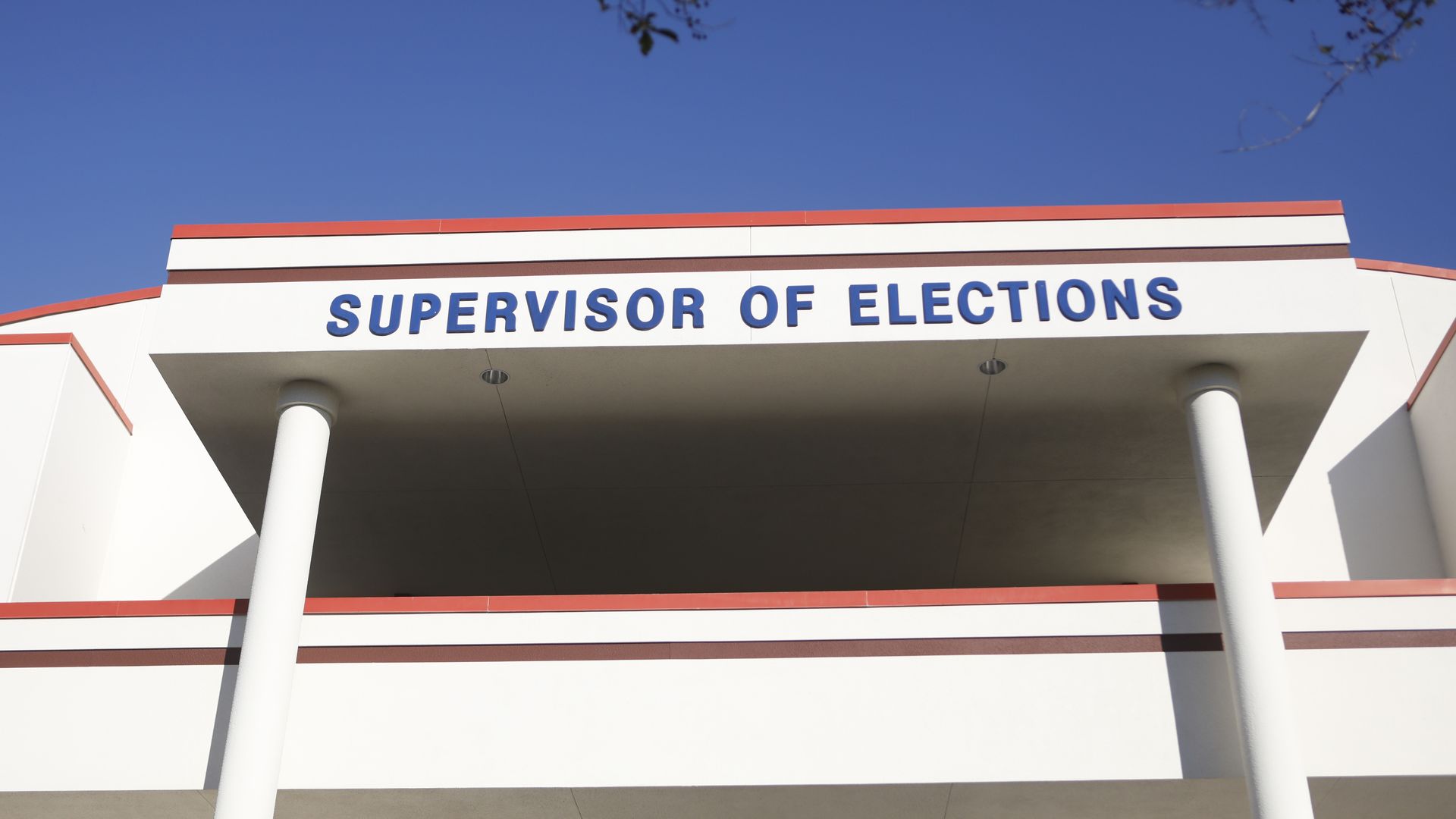 Blue sky above a white building with orange trim; a sign reads "SUPERVISOR OF ELECTIONS" in blue letters beneath a covered entrance supported by white columns.