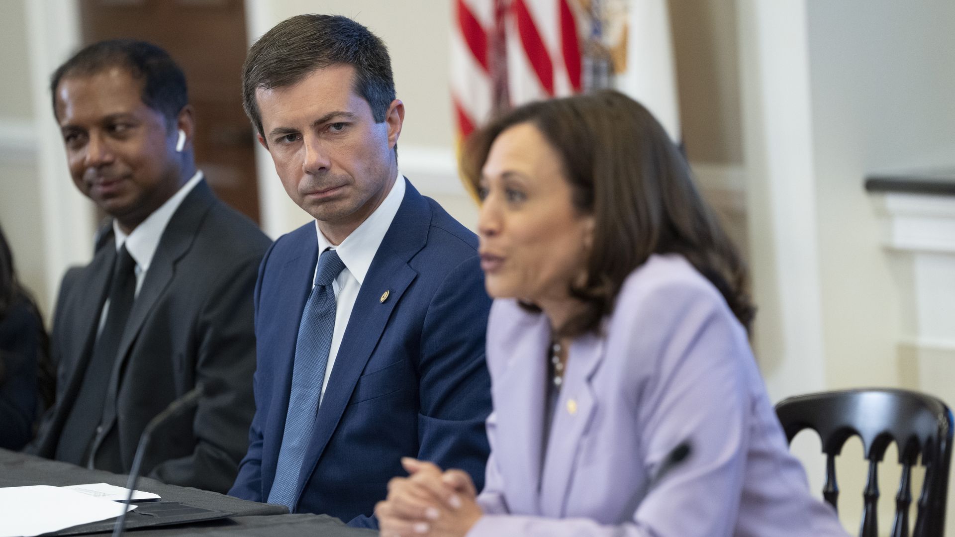 Pete Buttigieg, wearing a dark blue suit and tie, watches as Kamala Harris, wearing a light purple blazer, speaks into a microphone. The pair are sitting next to one another at a table.