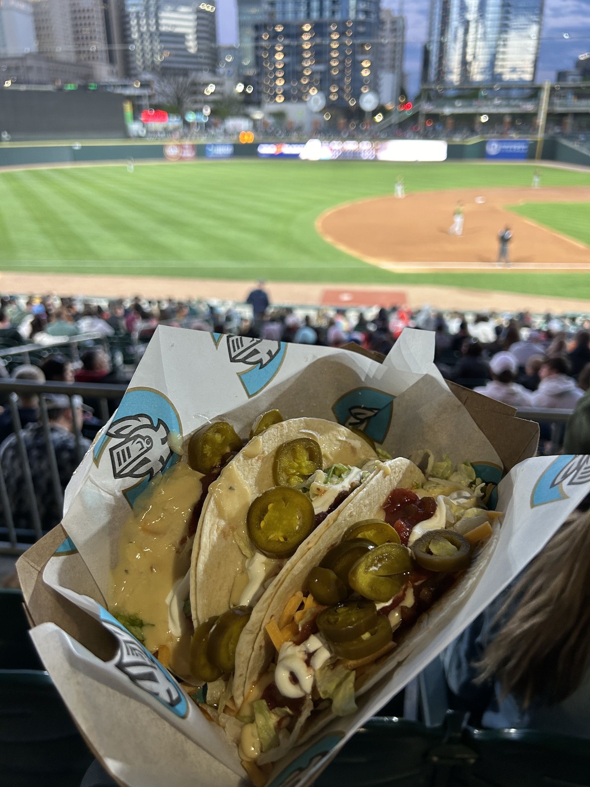 Three tacos on a plate, in front of a baseball stadium field.