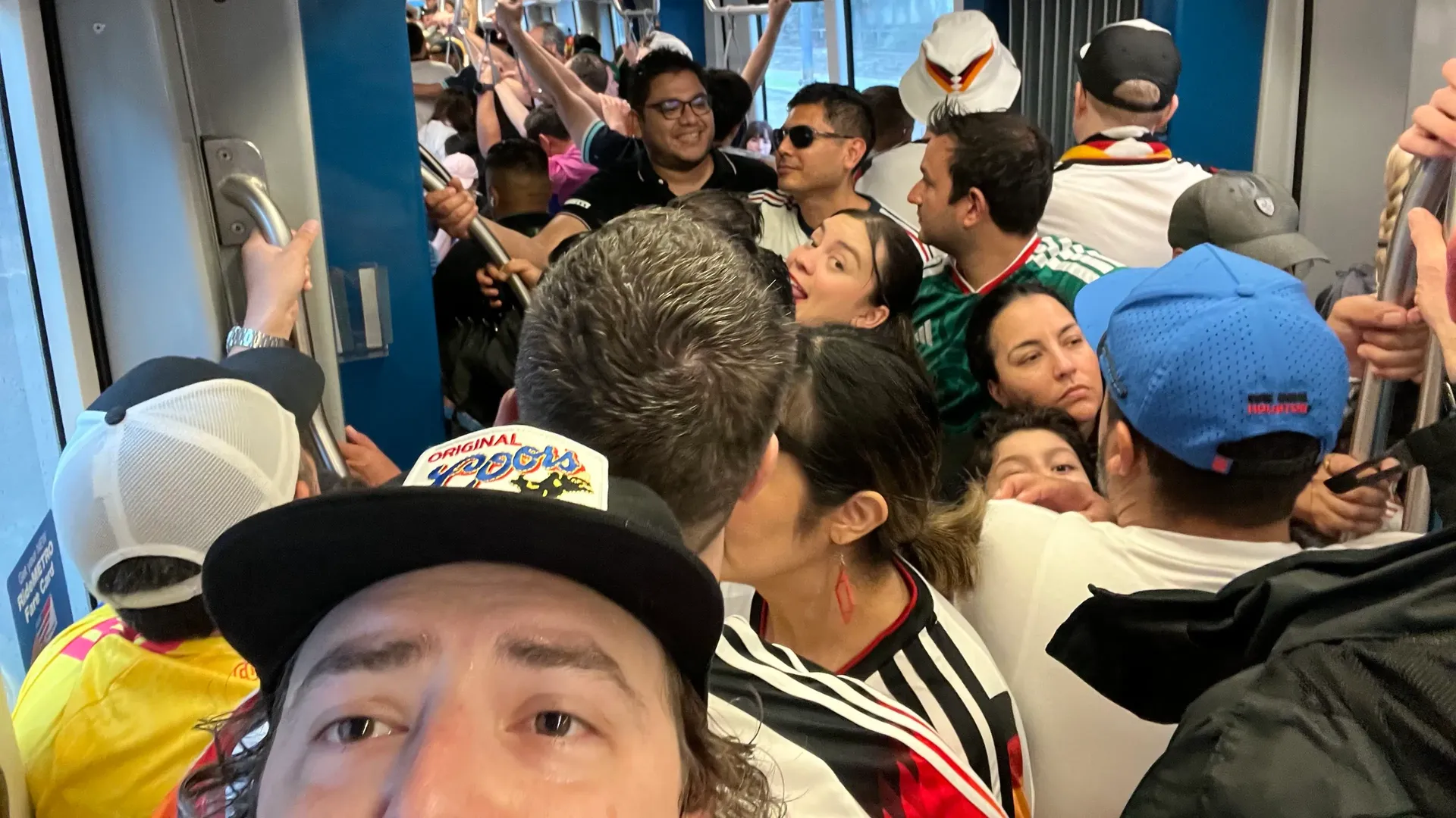 Overcrowded train car with standing passengers gripping rails and straps. A smiling man wearing sunglasses sits near the center; others wear colorful outfits. Blue interior walls.