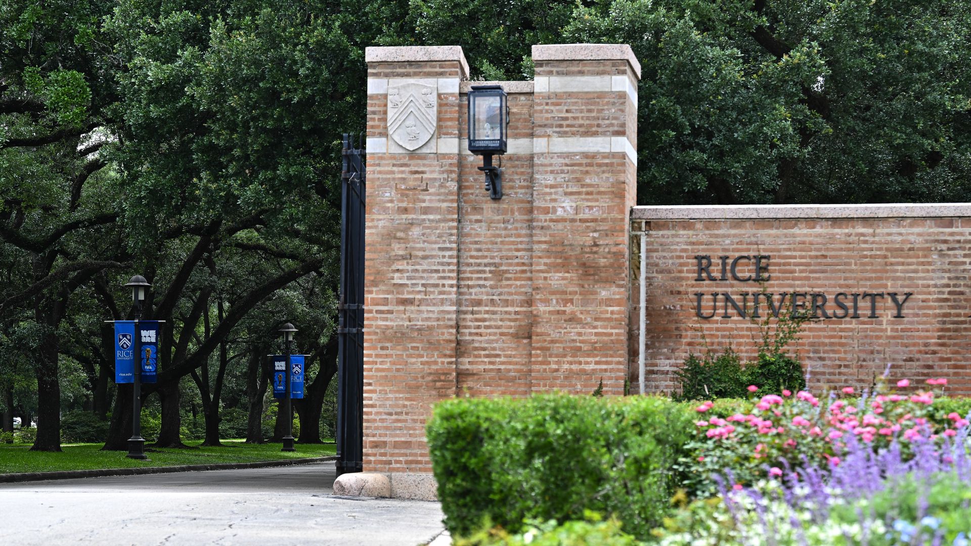 Photo of the entrance of college campus, with a brick gate with the words "Rice University." Greenery surrounding.