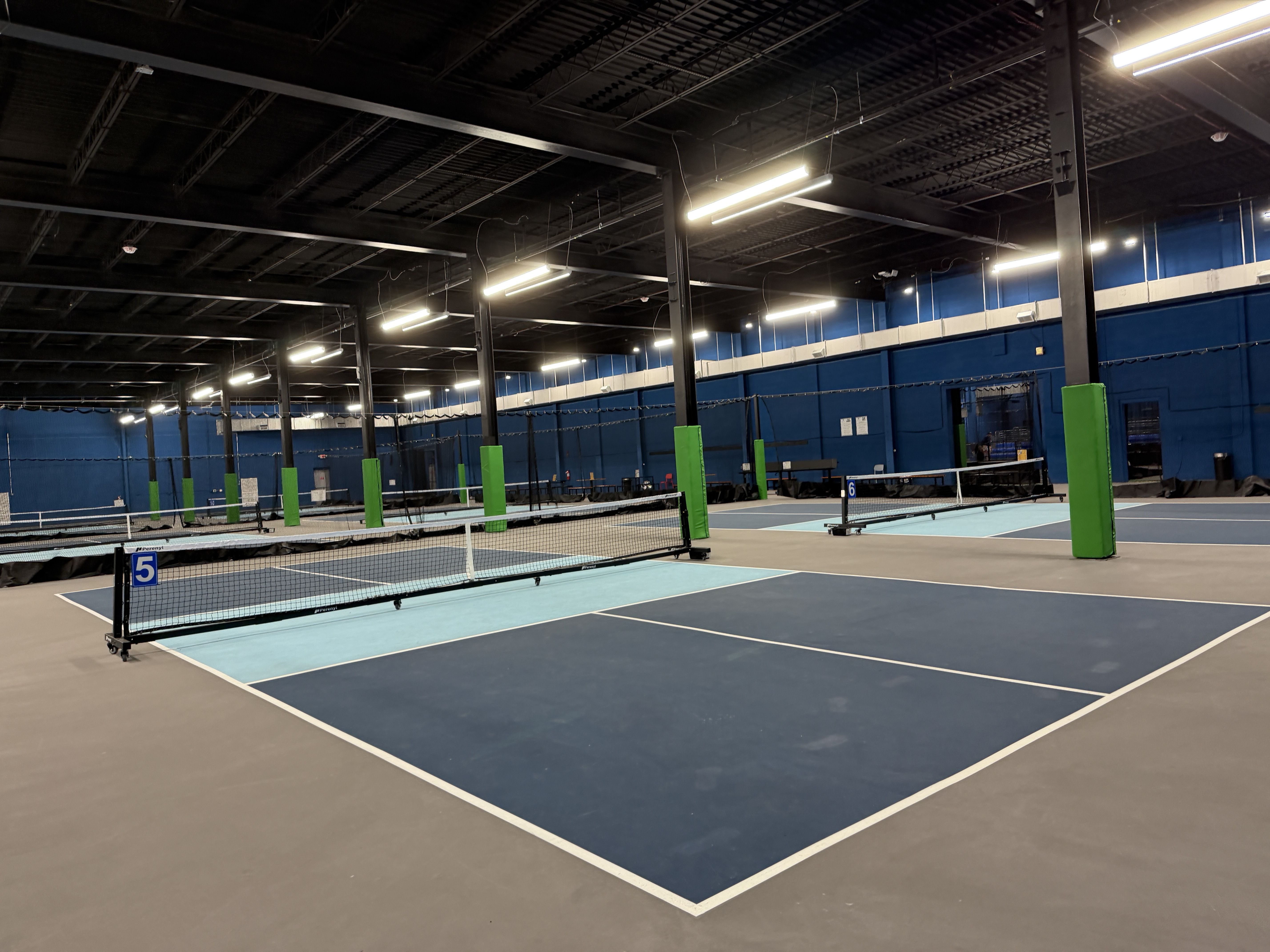 Interior of a large indoor tennis facility with multiple blue courts, white nets, and bright overhead lights. Black ceiling with metal beams; green padded columns along the court edges.