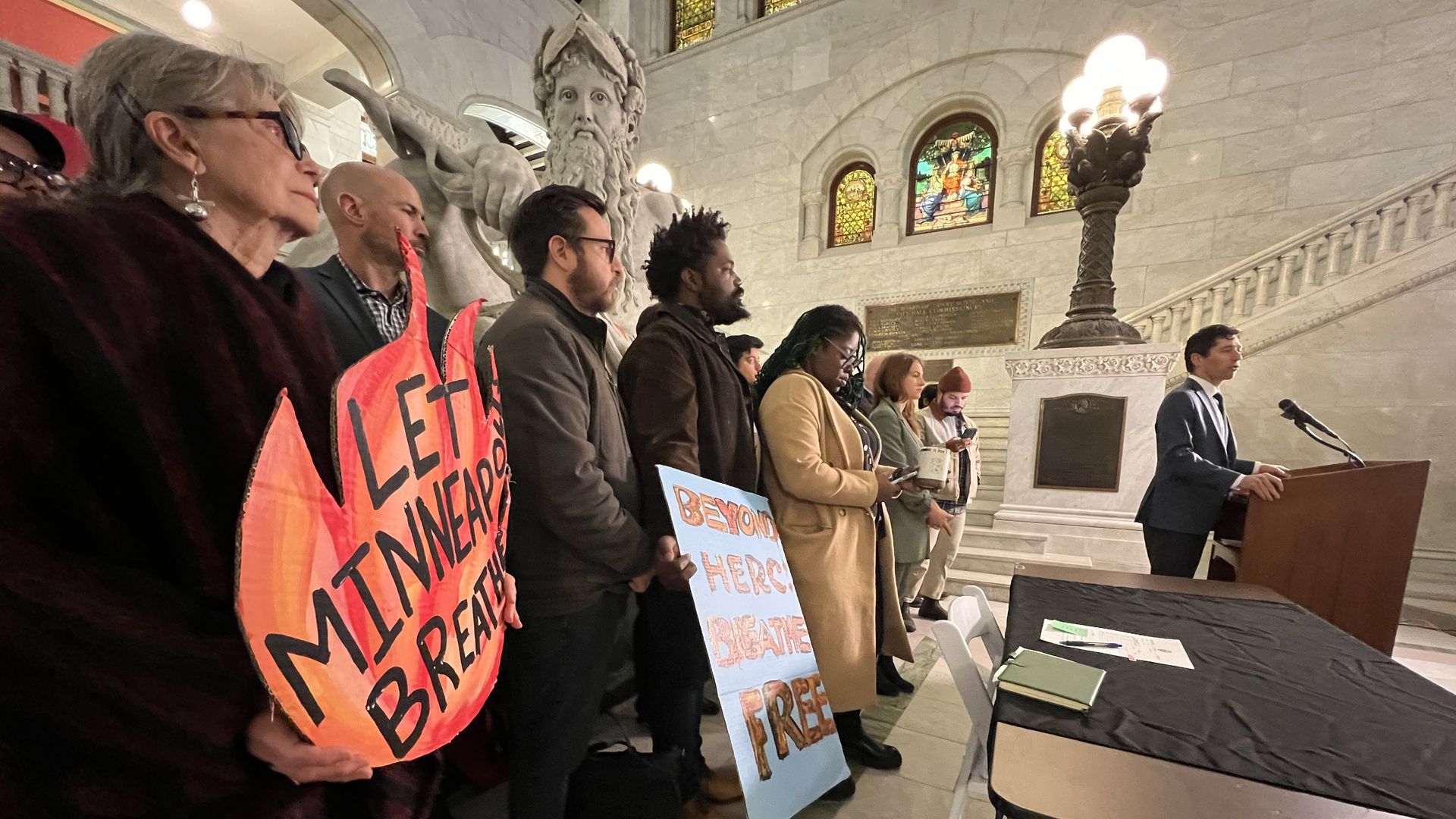 A woman holds up a sign in the shape of a flame reading "Let Minneapolis Breathe" in a large, marble hall