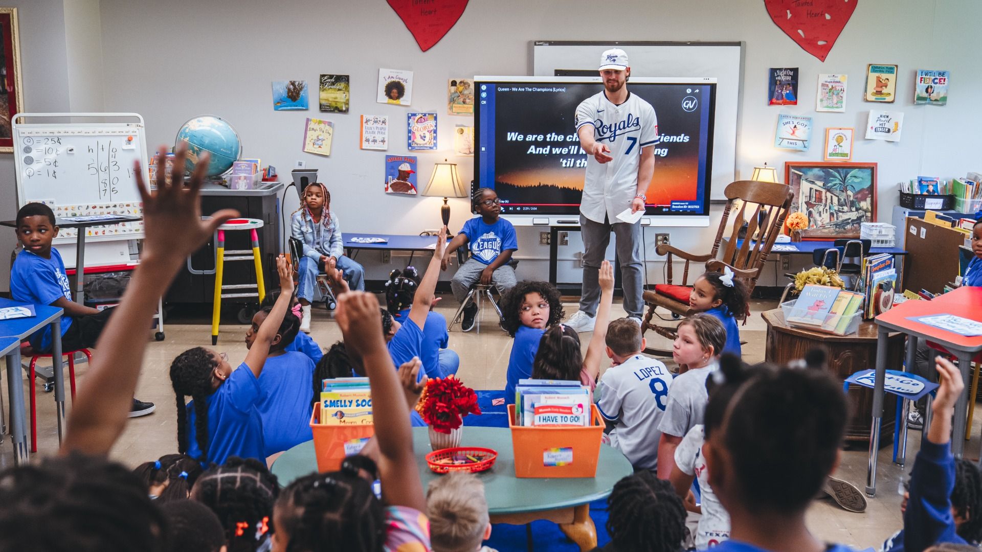 Bright classroom: a teacher in a Royals jersey stands before a screen with song lyrics as students sit and raise hands; posters, a globe, and books line the walls.