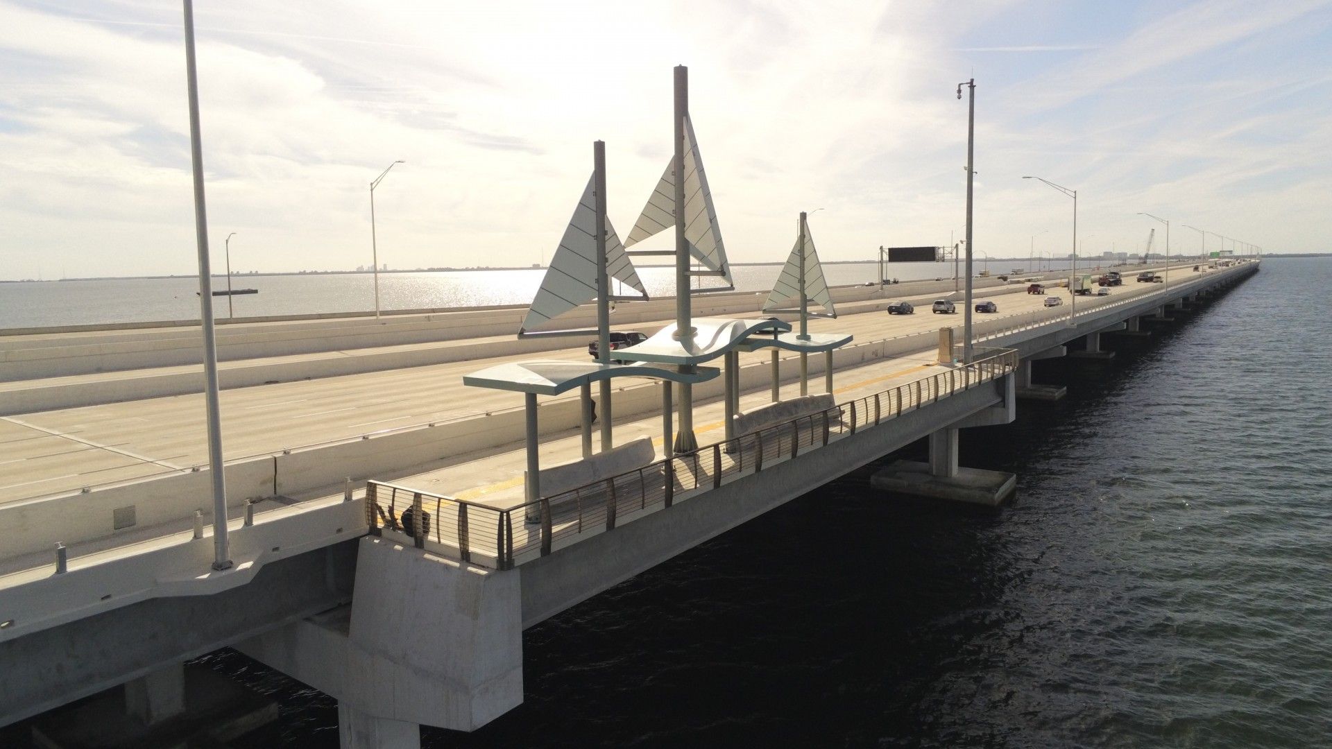 Bridge over water with a sidewalk and decorative sail sculptures along the railing; cars travel the roadway as the sun shines under a partly cloudy sky.