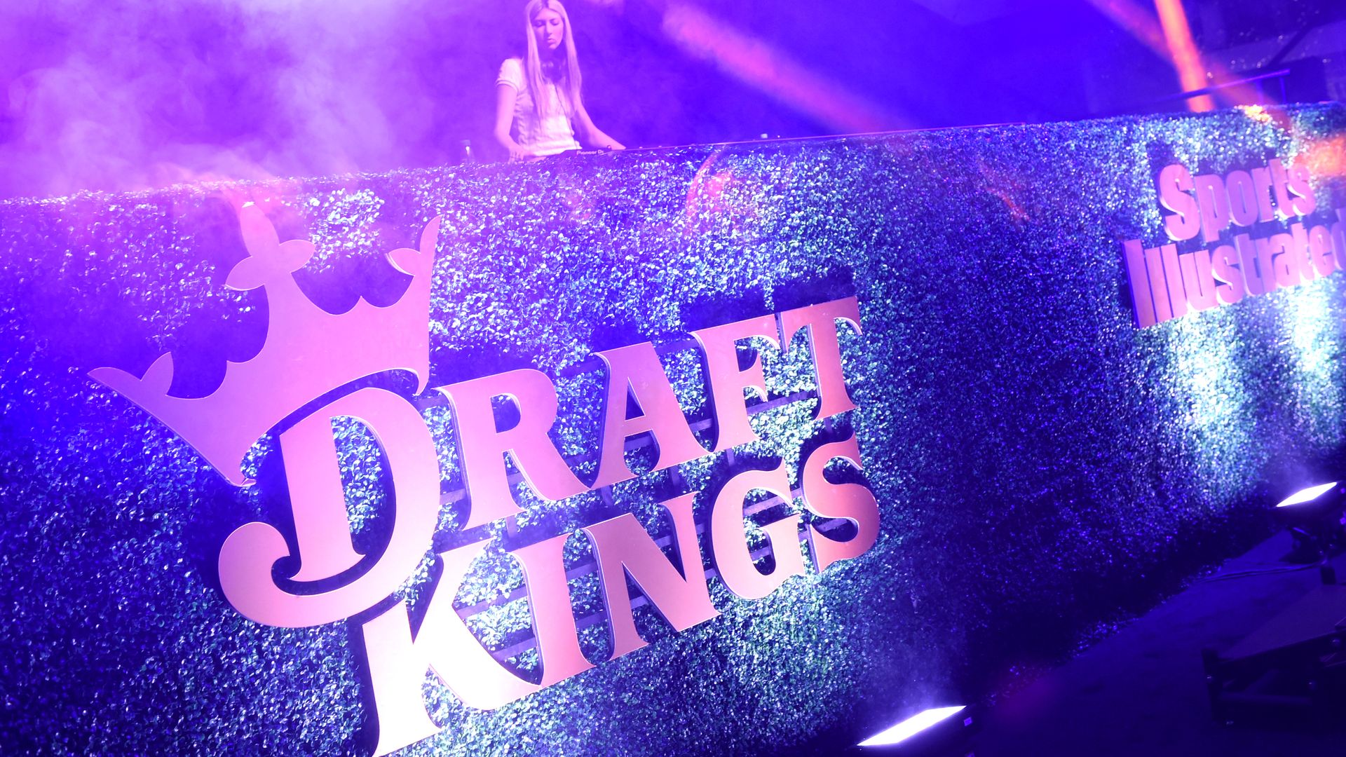 A female DJ performing behind a glittery blue wall with pink neon lights displaying "Draft Kings" and "Sports Illustrated" logos, surrounded by purple and pink stage lighting and smoke effects.