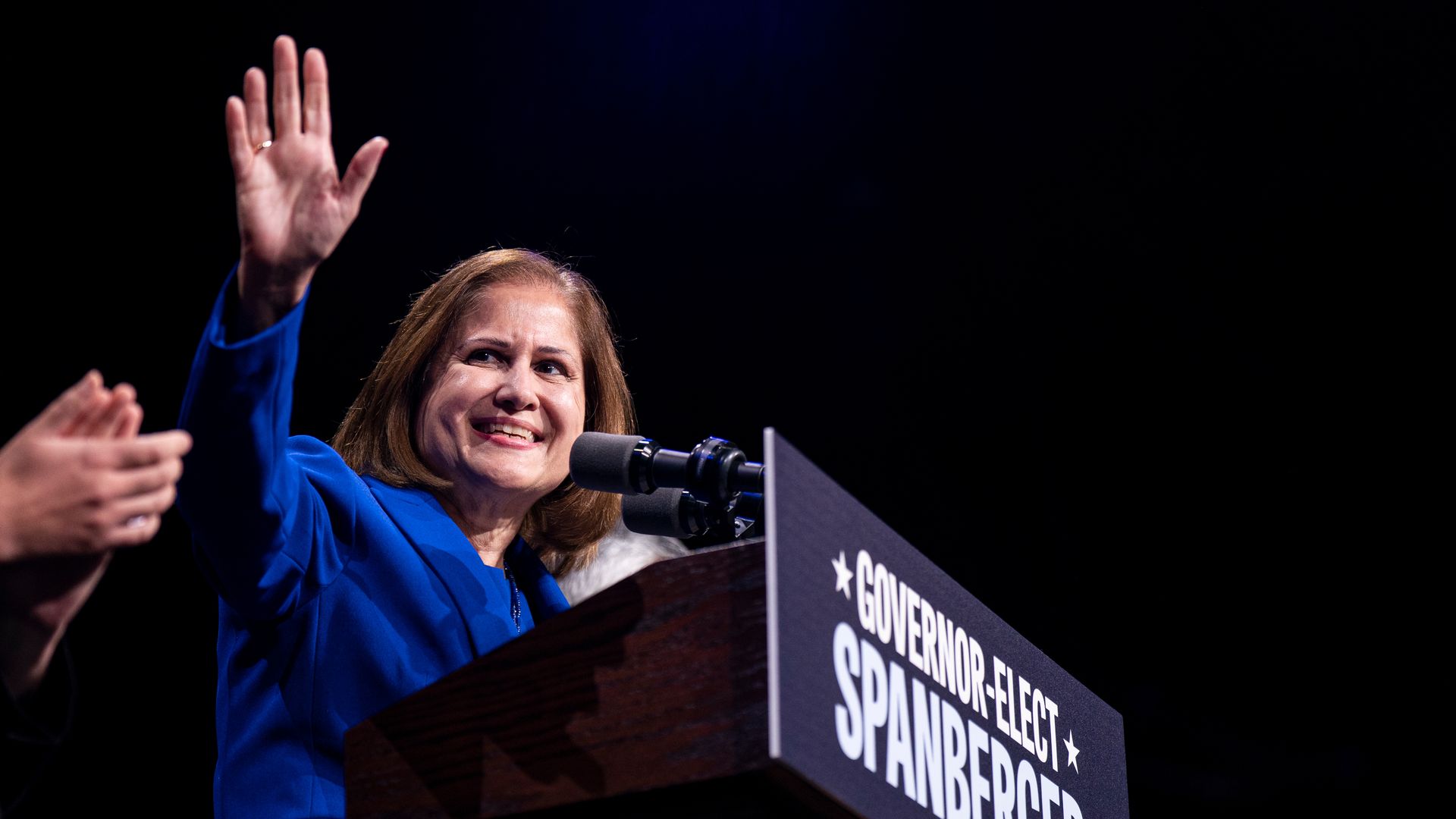 Ghazala Hashmi in a blue suit waving at the crowd from a podium