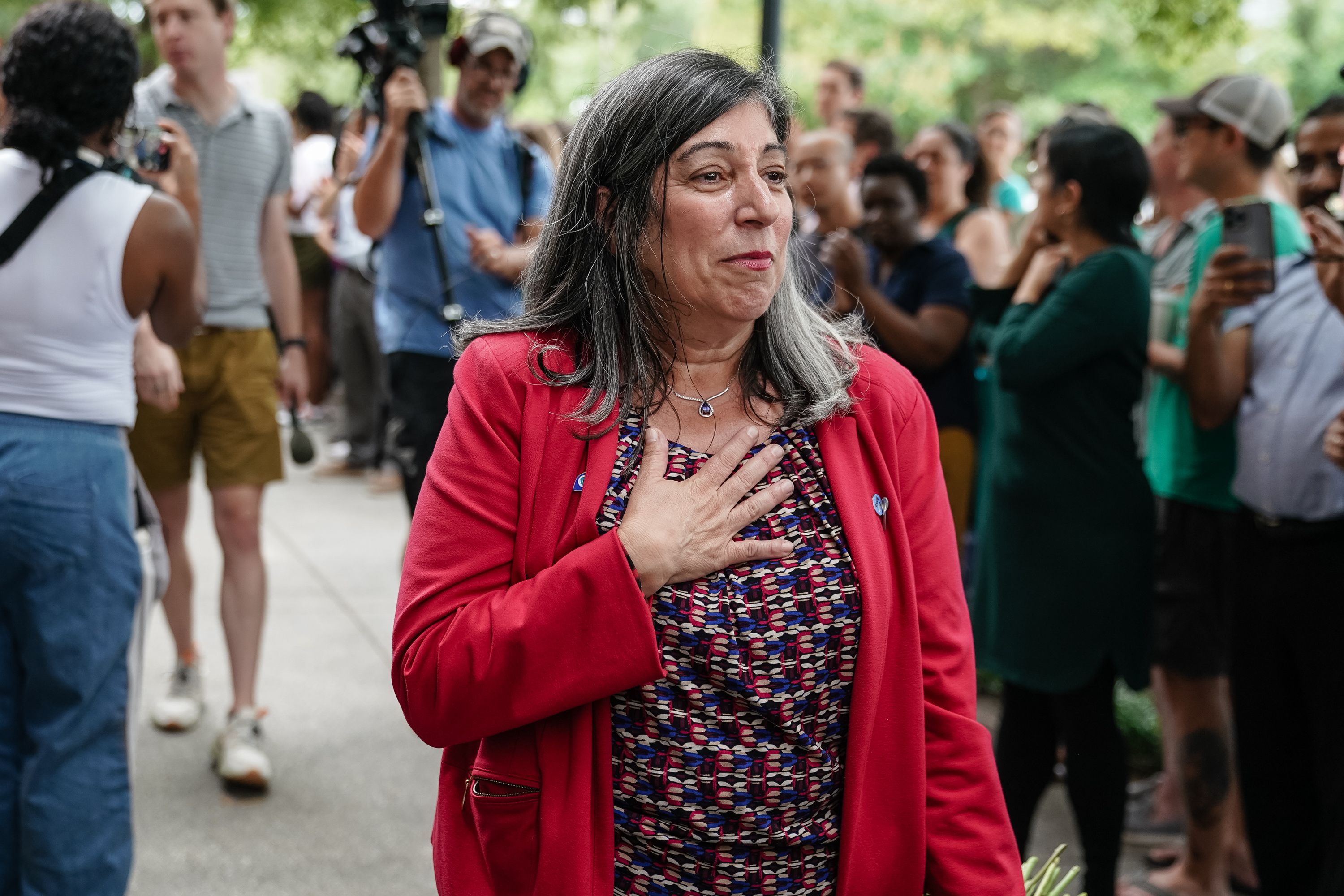 Smiling woman with gray hair in a red blazer and patterned dress holds hand to chest. Behind her, a diverse crowd stands outside, some taking photos and videos.