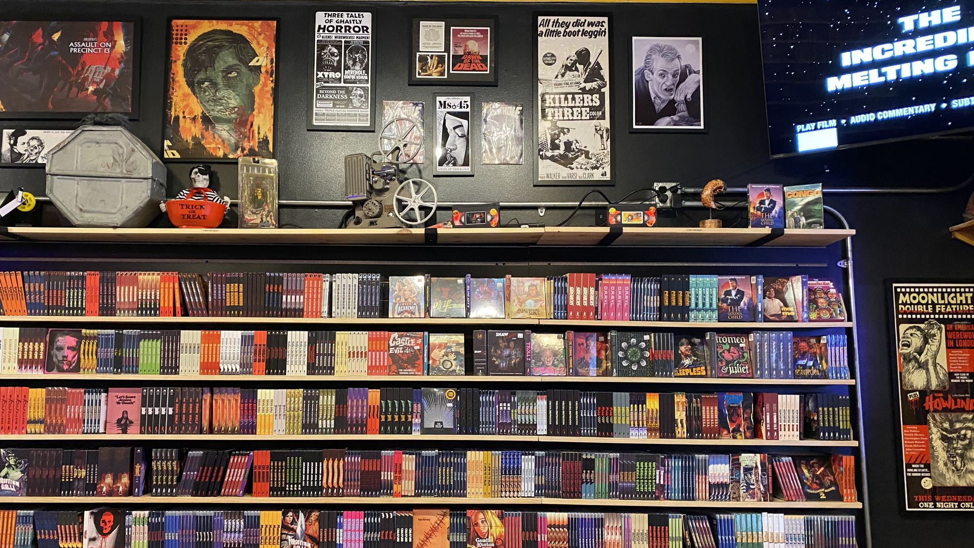 Wall with shelves filled with DVDs and VHS tapes, vintage horror movie posters and memorabilia above, a TV screen on the right showing a film title. Dark background with colorful covers.