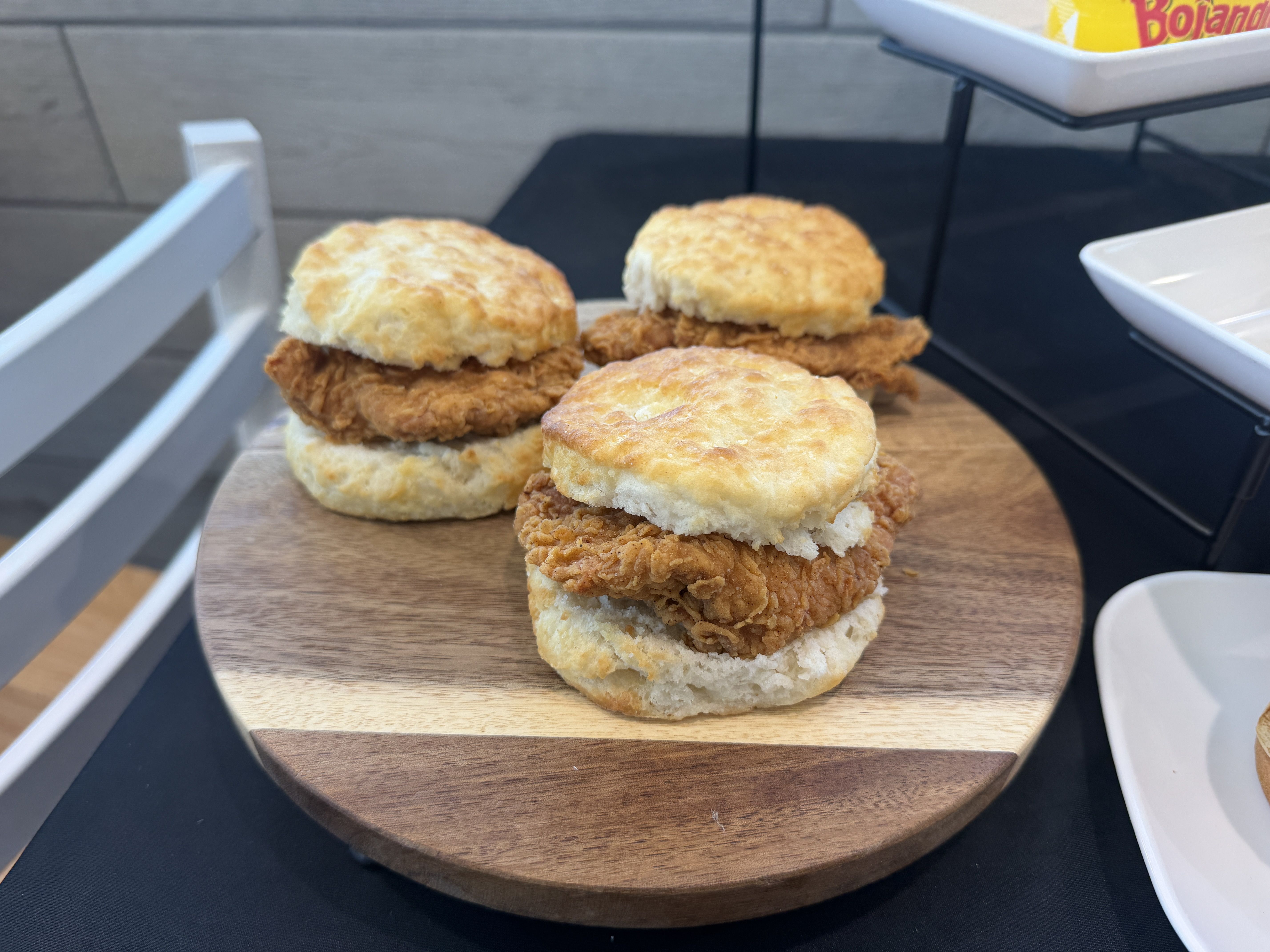 Three fried chicken sliders on a wooden board with flaky biscuits and crispy chicken, outdoors on a table, with a Bojangles bag visible in the background.