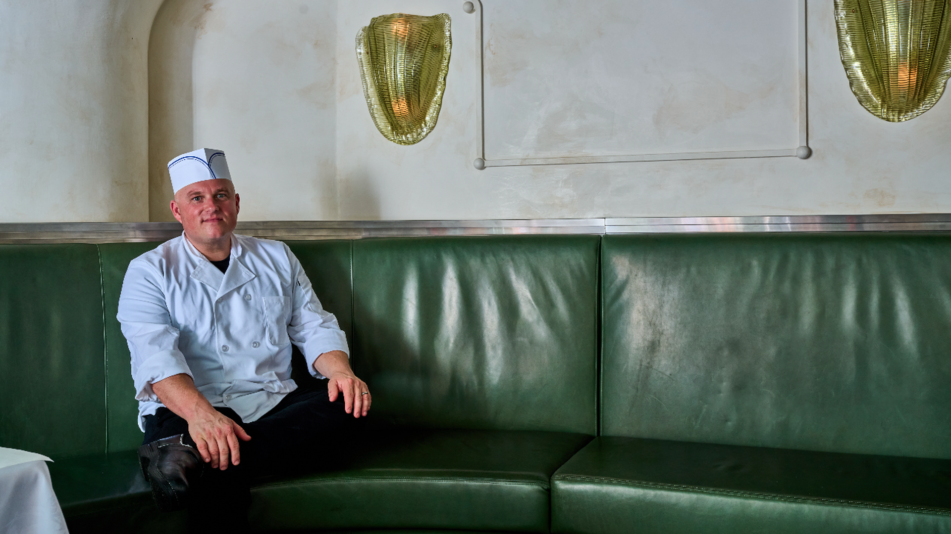 A chef in a white uniform and hat sits on a dark green leather couch in a retro diner-style room with pale walls and gold wall sconces.