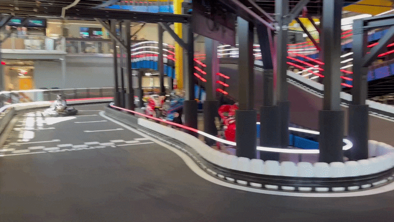 A go-kart racing across an indoor track with red and white lights flashing in the background.