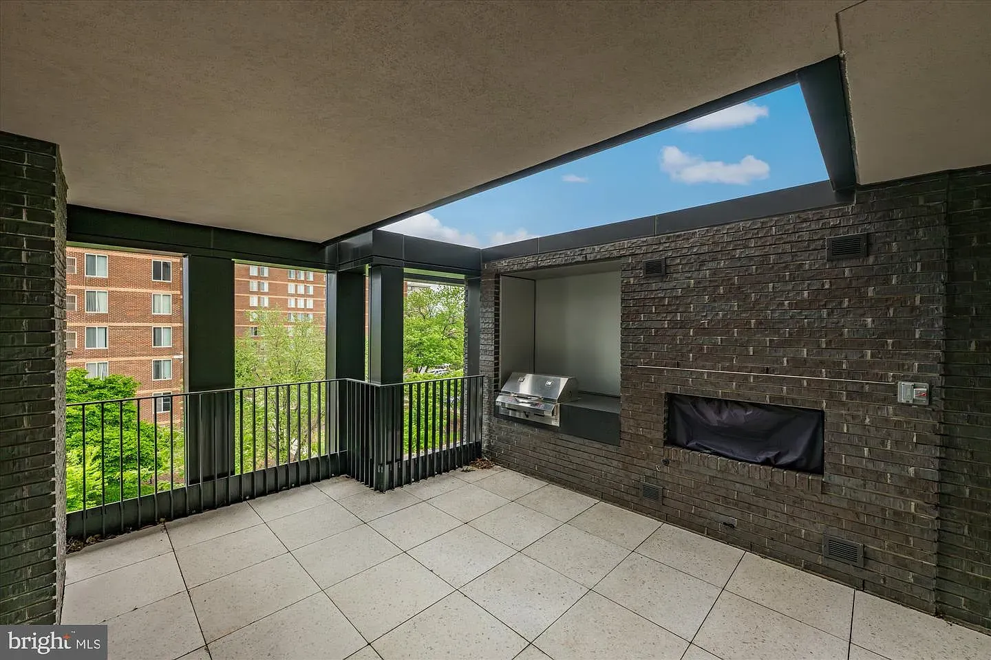 Outdoor balcony with dark brick walls, light tile floor, and a black railing. A stainless steel grill sits in a recessed wall, beside a rectangular fireplace; view of trees and a red brick building.