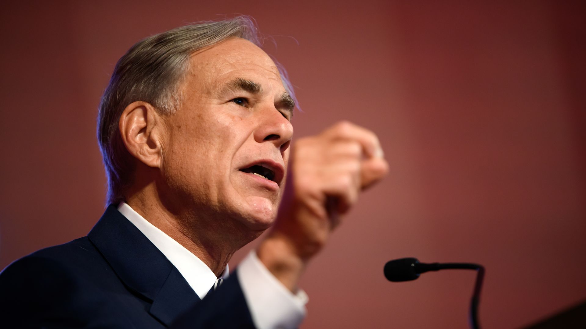 Gov. Greg Abbott, with gray hair in a dark suit speaking passionately at a microphone against a reddish background, gesturing with his right hand raised.