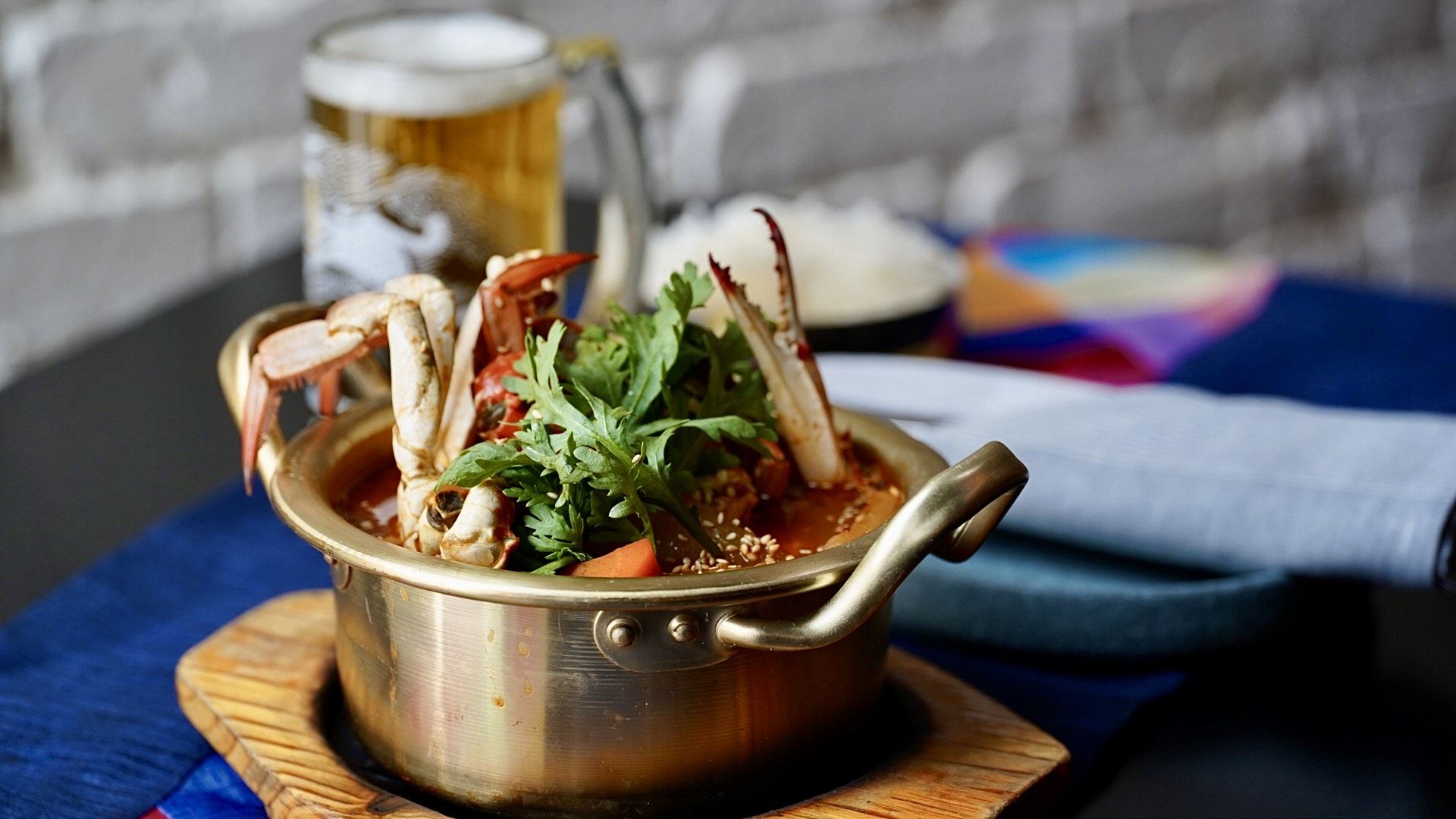 Brass pot filled with crab legs, green herbs, and red broth on wooden board, with a glass of beer, bowl of white rice, and folded napkin in the background on a blue tablecloth.