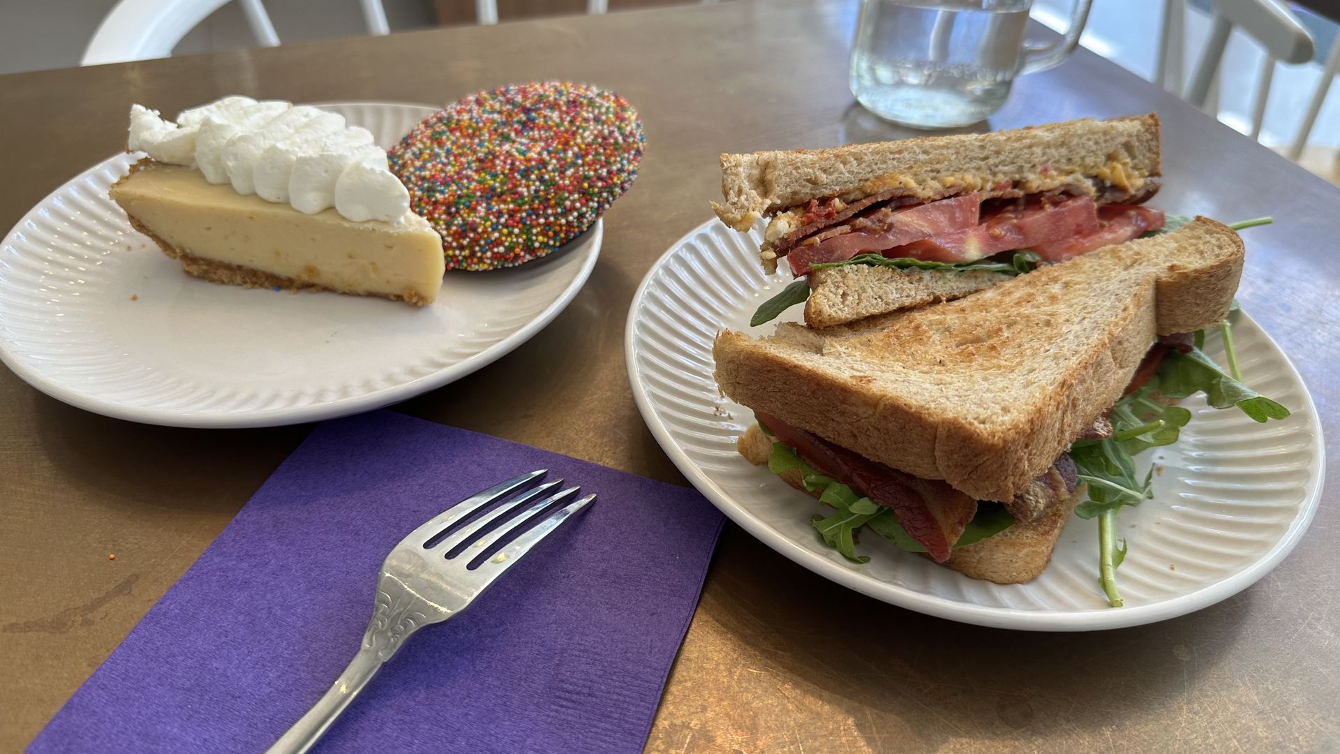 A fork sits on a purple napkin next to two plates on a table. One plate has a slice of key lime pie and a cookie, and another plate has a BLT sandwich.