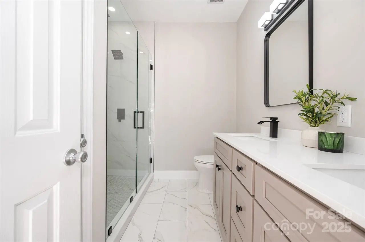 Modern bathroom with white marble floor, beige walls, glass shower enclosure, white toilet, long beige vanity with black knobs, black faucet, rectangular mirror, and green plant on countertop.