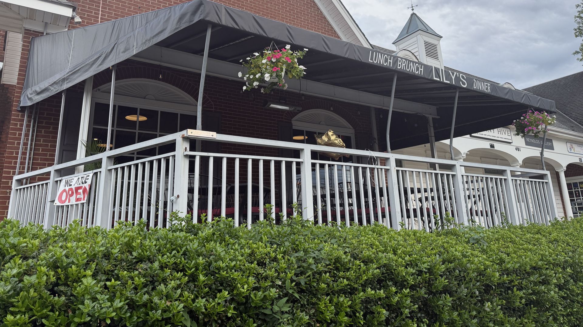 Brick storefront with a gray canopy over outdoor seating. The awning shows "LUNCH BRUNCH" and "LILY'S DINNER." A hanging flower basket and white railing with green hedges before a cloudy sky.