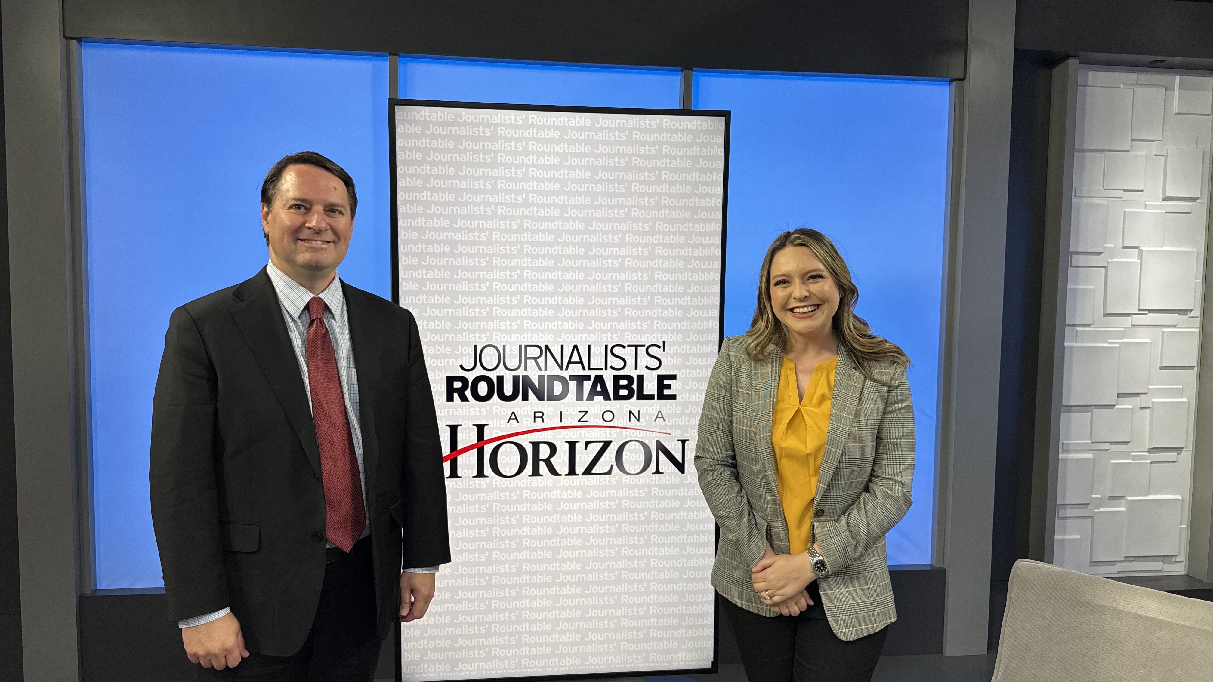 Two professionally dressed hosts stand beside a gray banner reading "Journalists' Roundtable" against a blue studio backdrop.