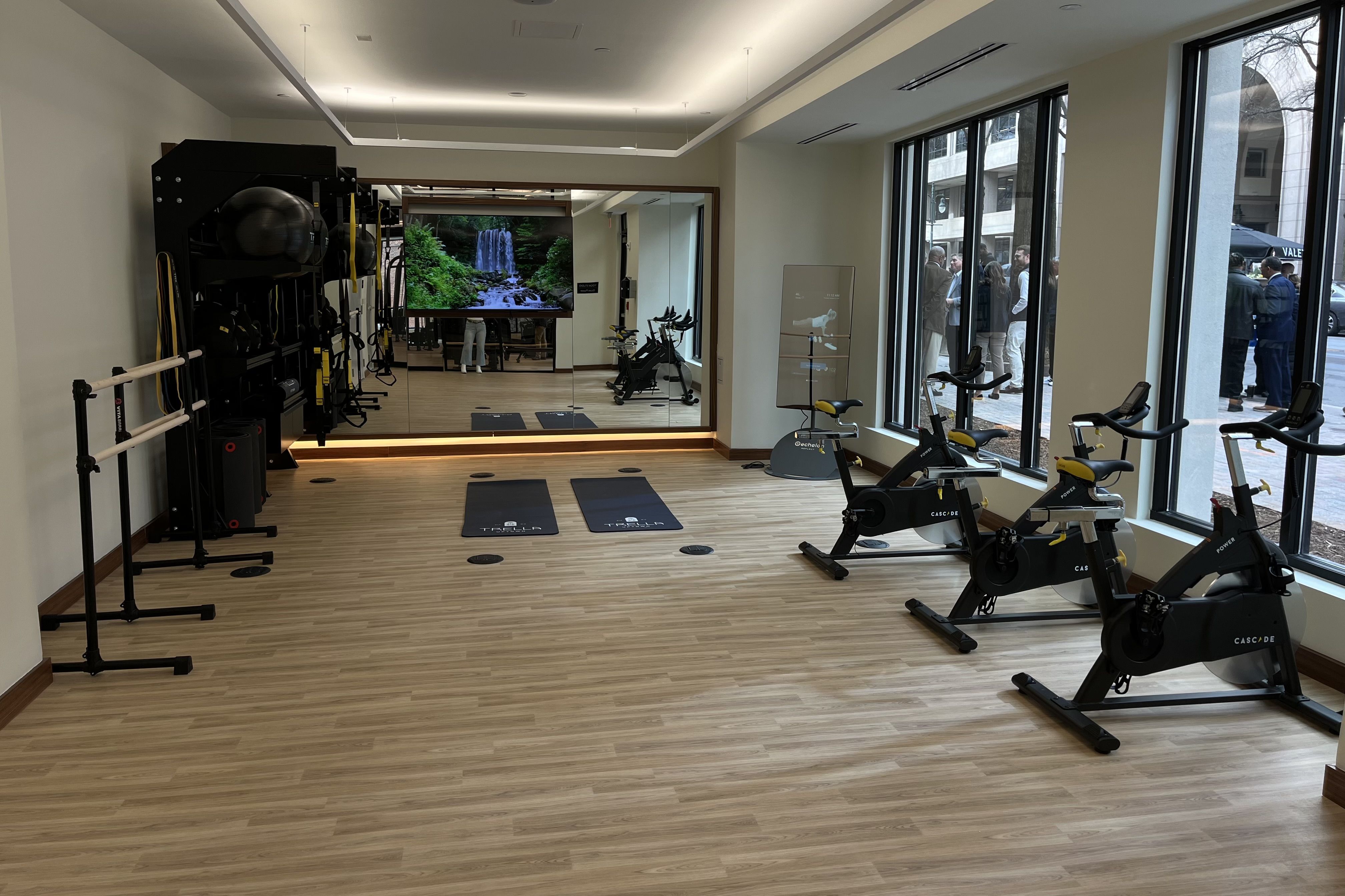 Bright gym with wooden floor, three black and yellow stationary bikes by large windows, two black exercise mats, wall-mounted workout equipment, and a screen showing a waterfall.
