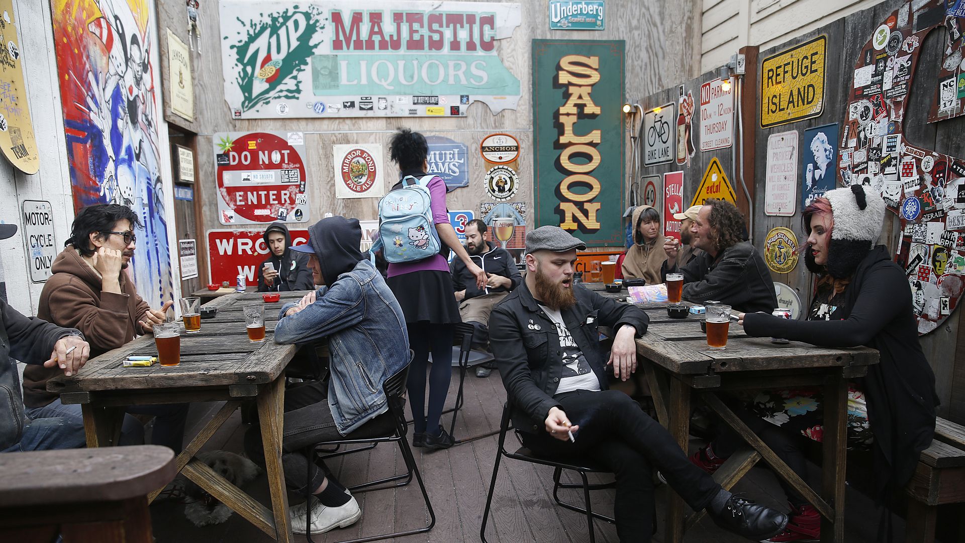 Outdoor rustic bar with wooden picnic tables and walls covered in vintage signs. Friends chat and drink beer; a server walks by with a light blue backpack. Colorful murals surround the scene.