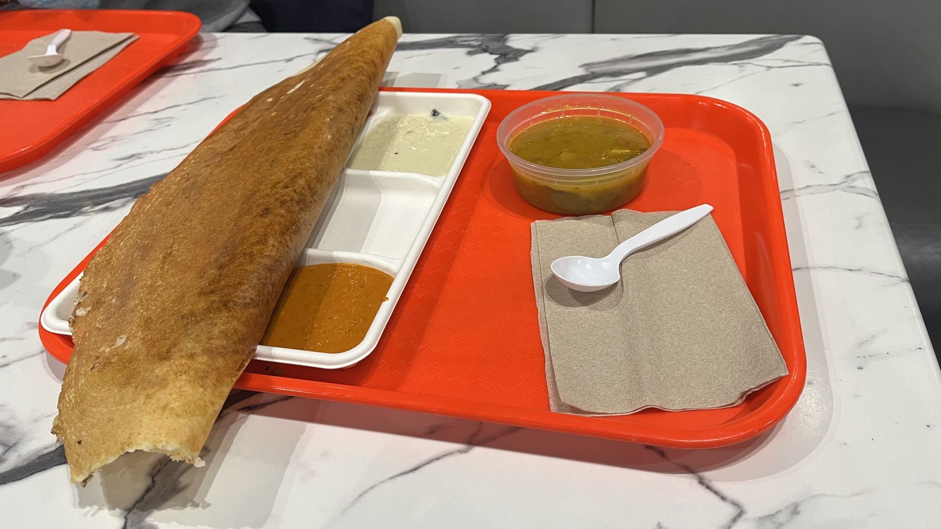 Large brown dosa on a white divided plate with reddish-orange and white chutneys, green sambar in a plastic cup, spoon on brown napkins, all on an orange tray on a marble table.