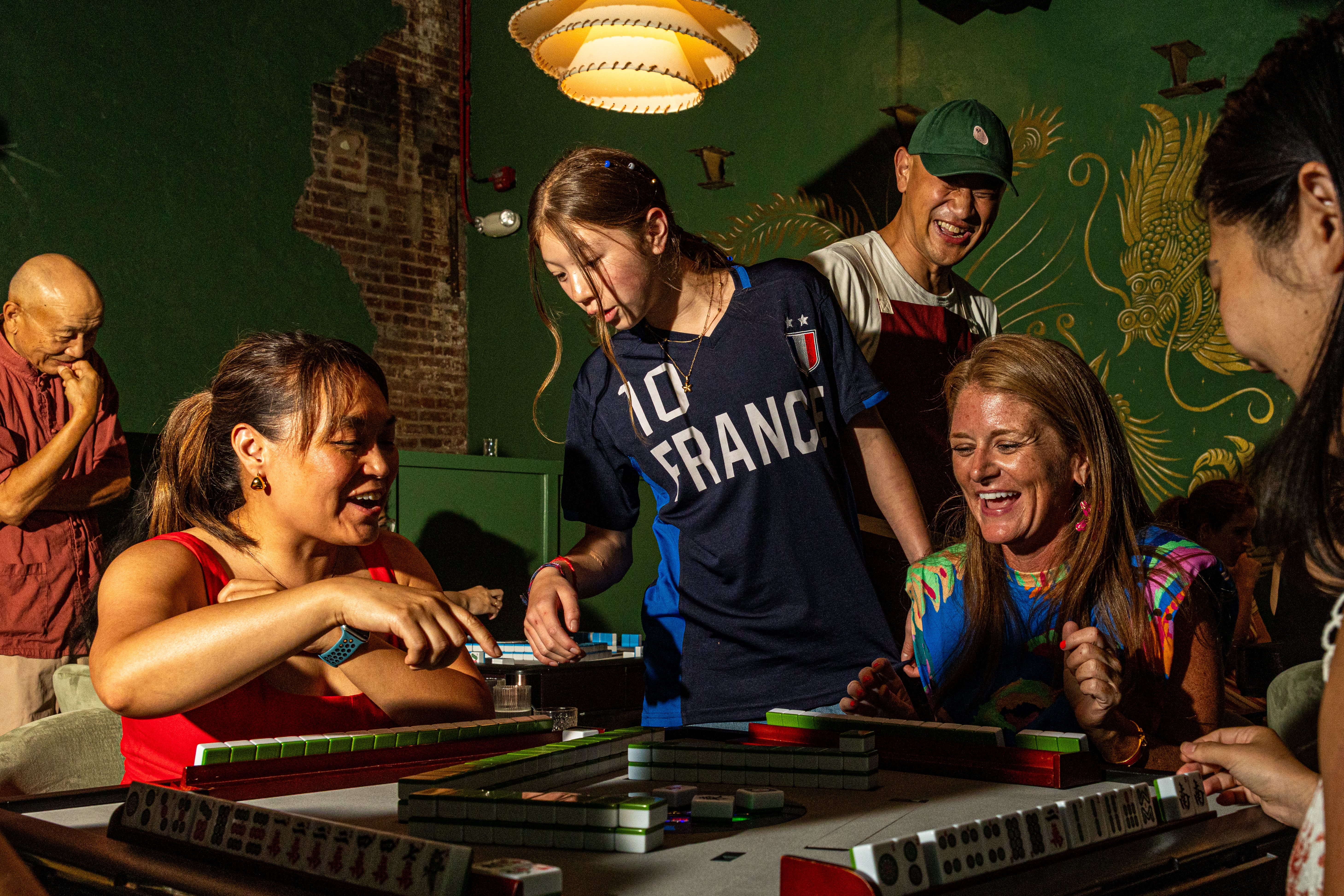 Group of people joyfully playing mahjong indoors with green walls and golden dragon mural, lit by warm overhead lamp in a lively setting.