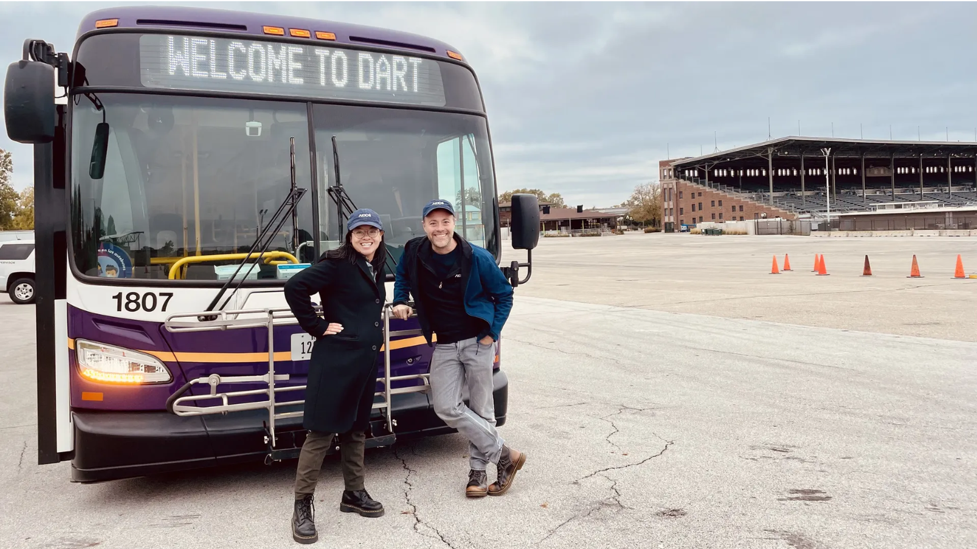 Two people smiling, wearing blue hats, standing in front of a purple and white bus with "WELCOME TO DART" displayed on its screen, in a large open area near a stadium.