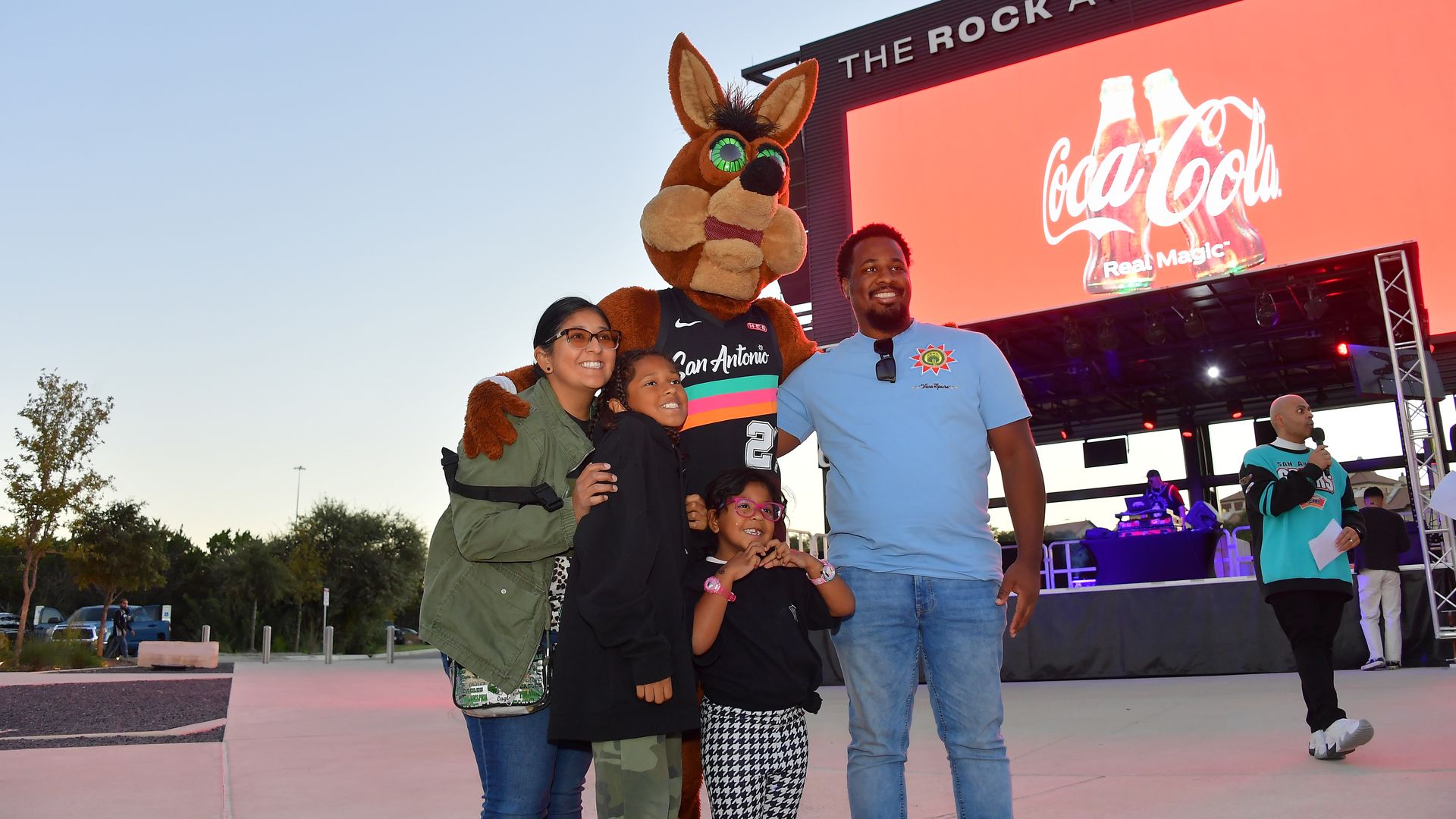 A family of four poses with the Spurs coyote mascot, who is wearing a San Antonio jersey. The photo is outdoors at dusk, near a stage called "The Rock at La Cantera" with a large Coca-Cola ad.