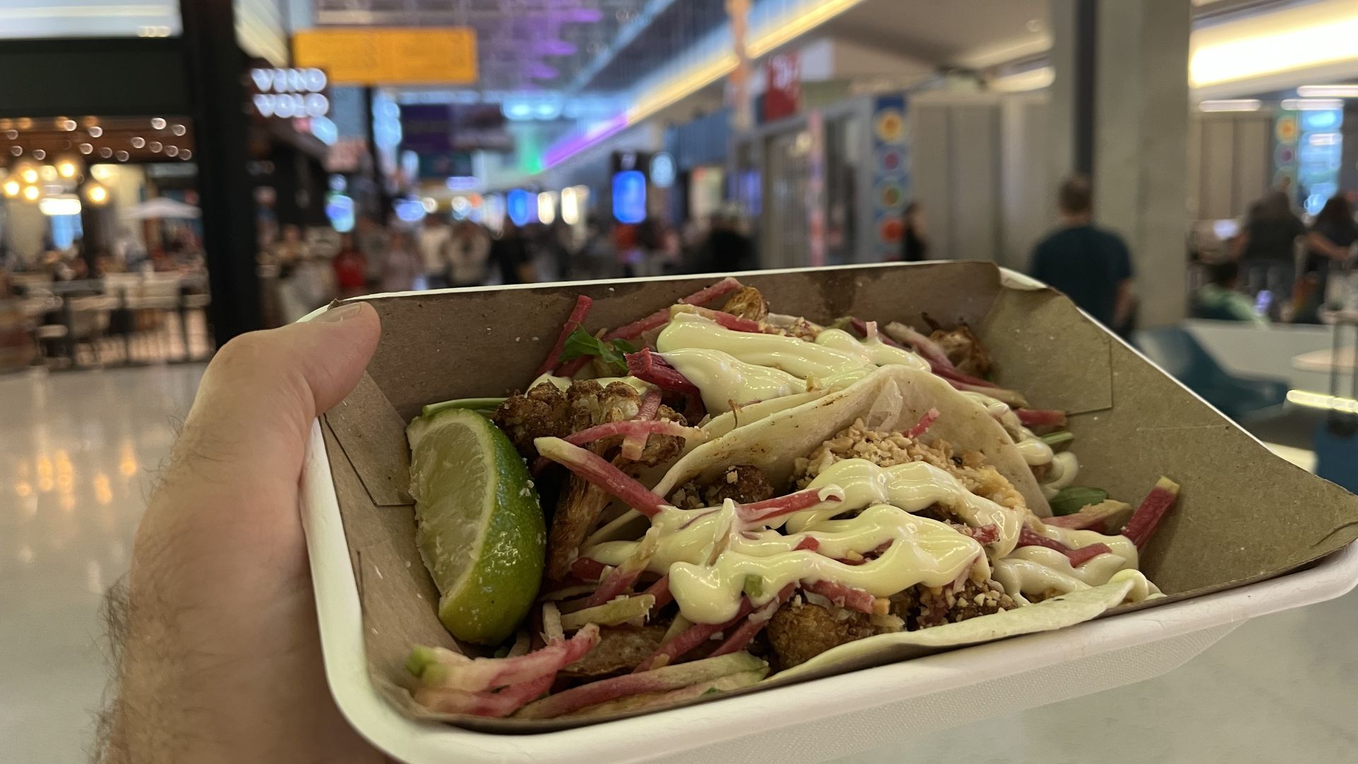 Hand holding a cardboard tray with two tacos topped with sauce, shredded vegetables, and a lime wedge, inside an airport.