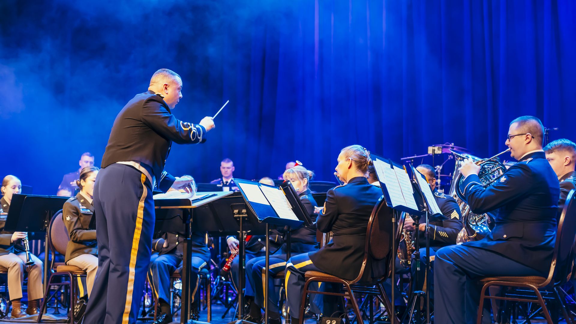 Conductor in a blue uniform with gold stripes directs a seated military band on a smoky blue-lit stage; brass players and sheet music stands are visible.